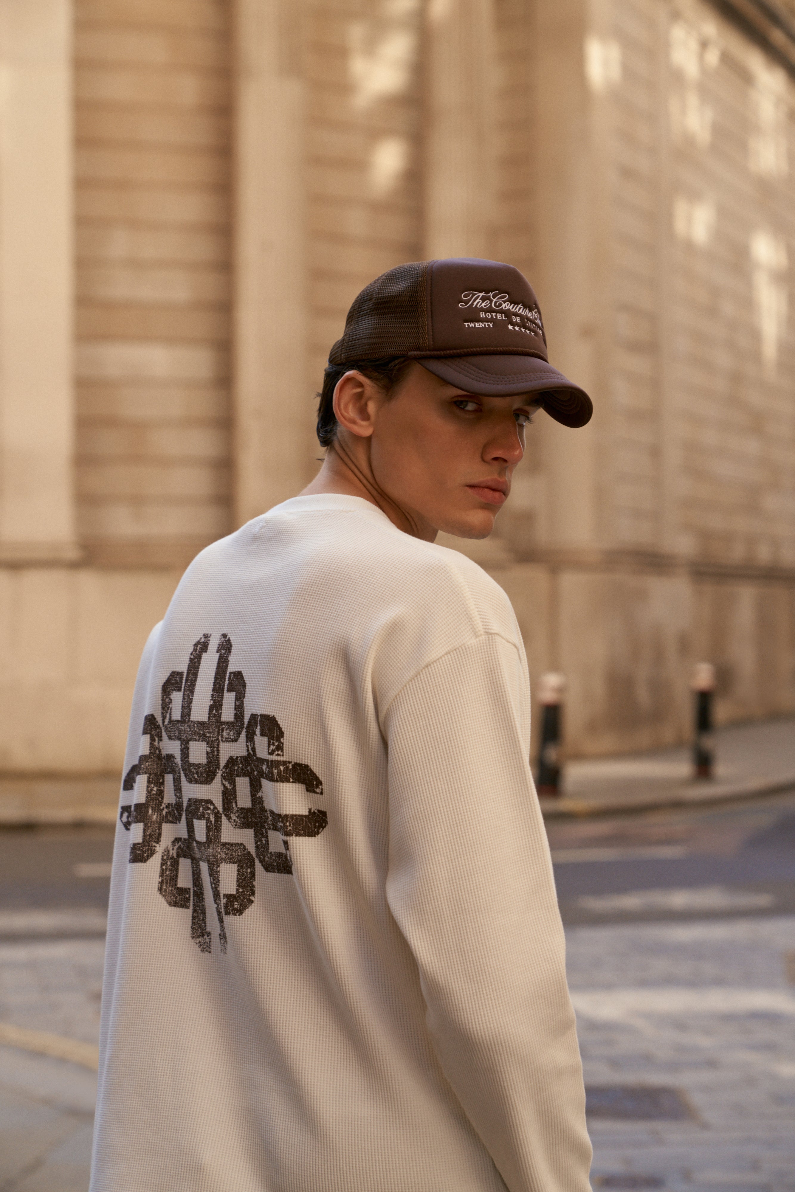 Person wearing a white t-shirt with black text on the back, standing against a beige wall.