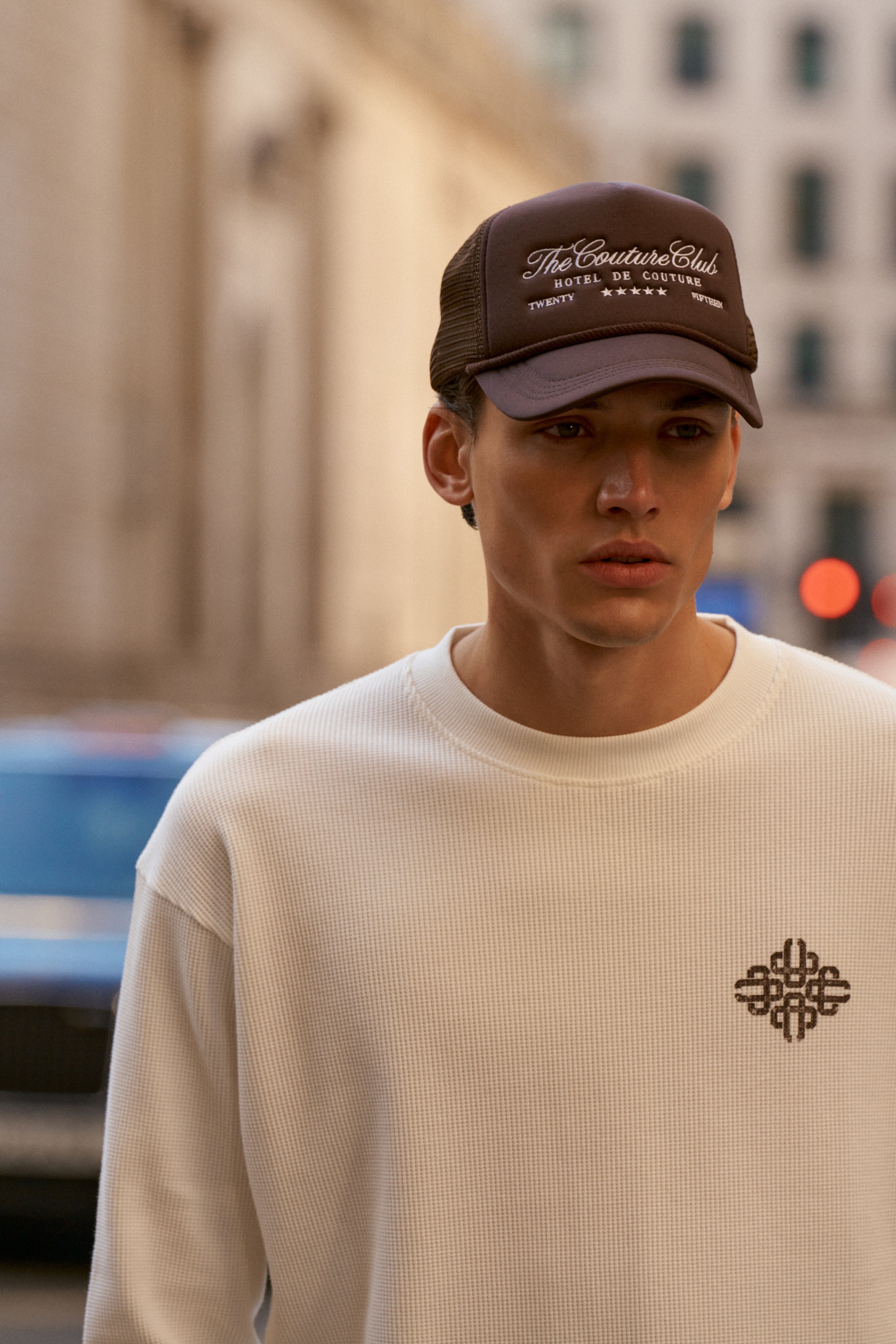 Man wearing a beige t-shirt with a logo and a dark cap in an urban setting