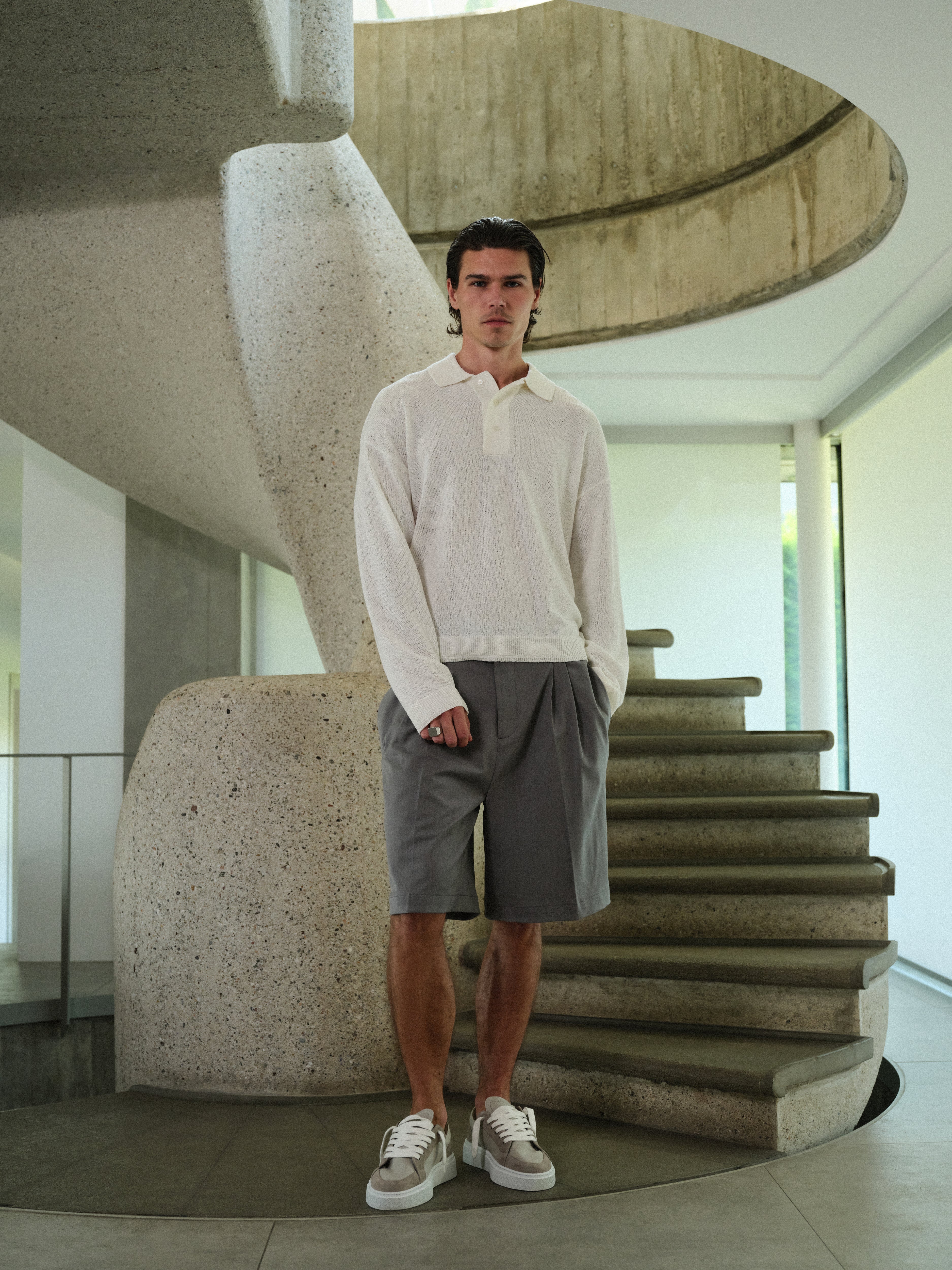 Man wearing grey tailored shorts, white long-sleeved polo shirt, standing in front of a modern staircase with spiral design.