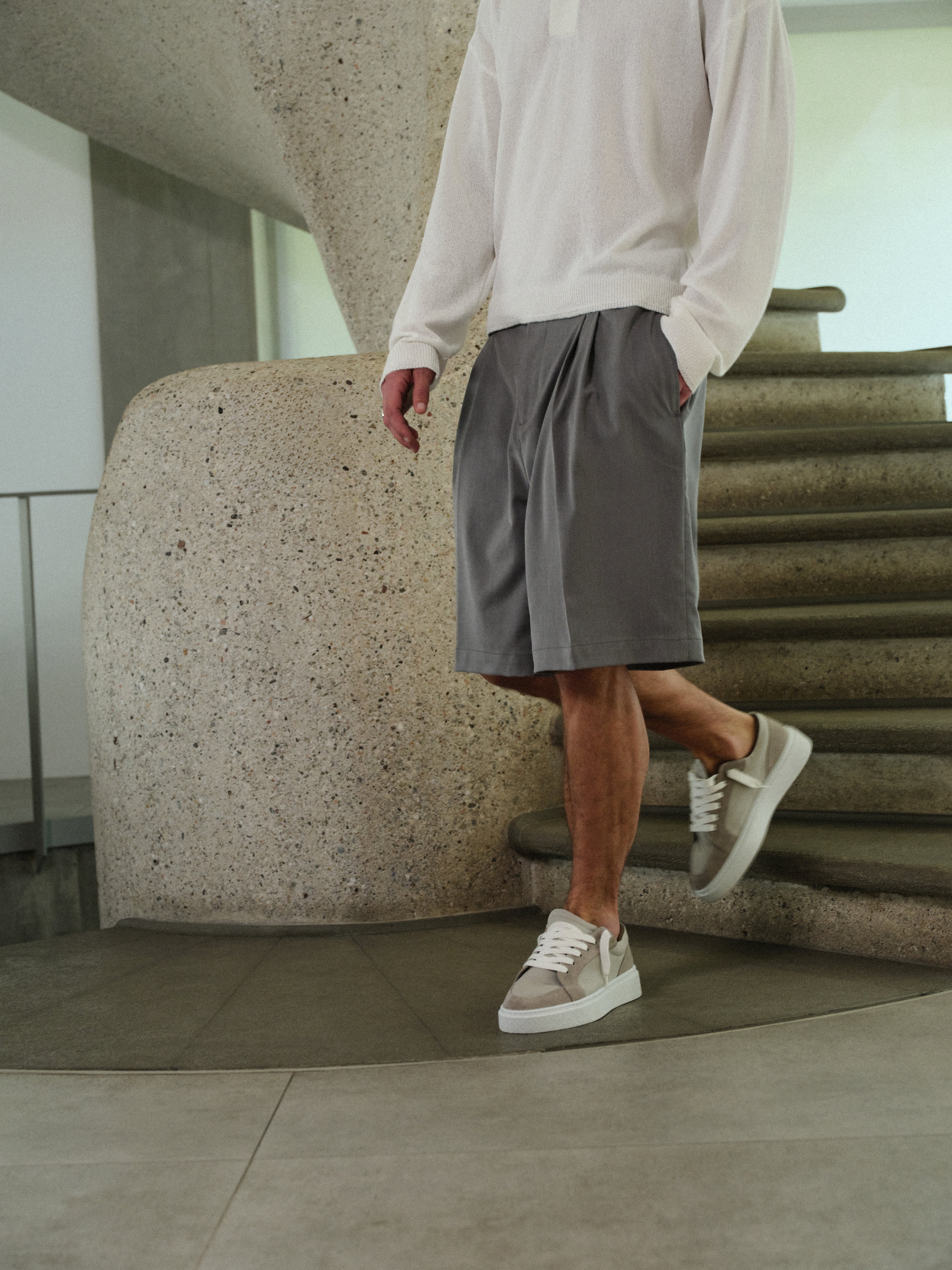Man wearing grey shorts and white sneakers standing in a modern indoor setting.