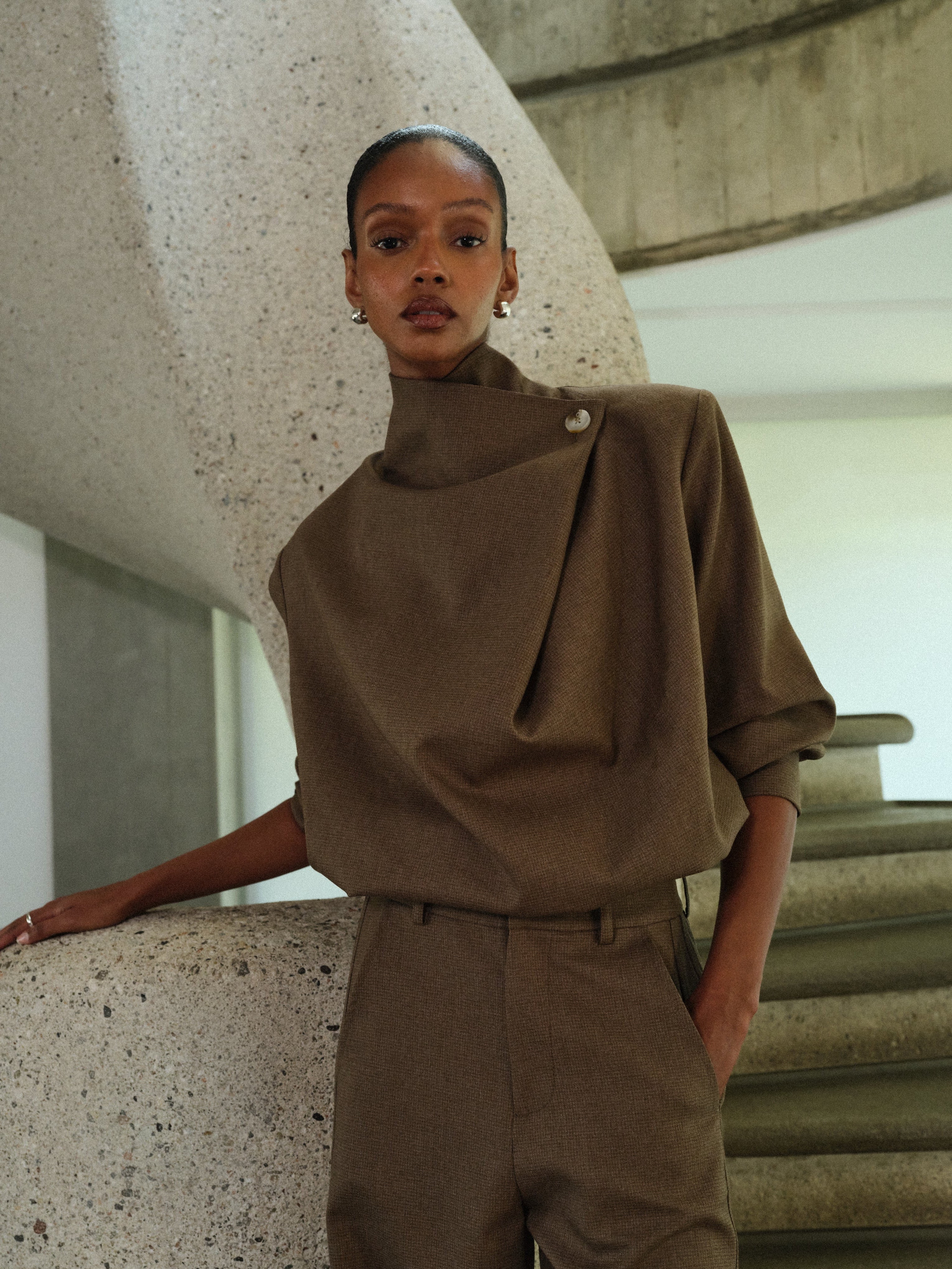 Person wearing a brown matching tailored outfit standing against a concrete wall.