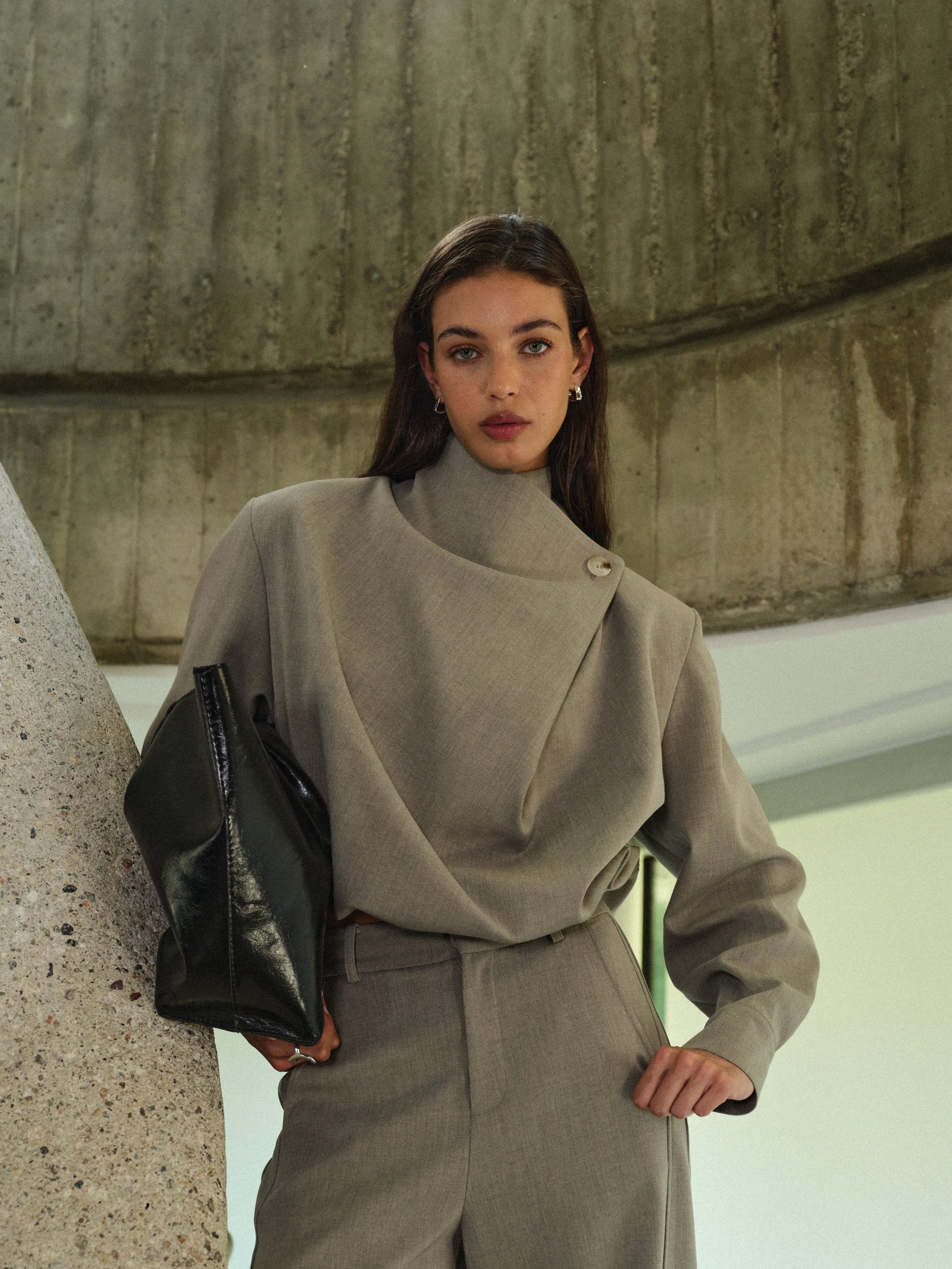 Woman in a grey matching set holding a black bag against a concrete wall.