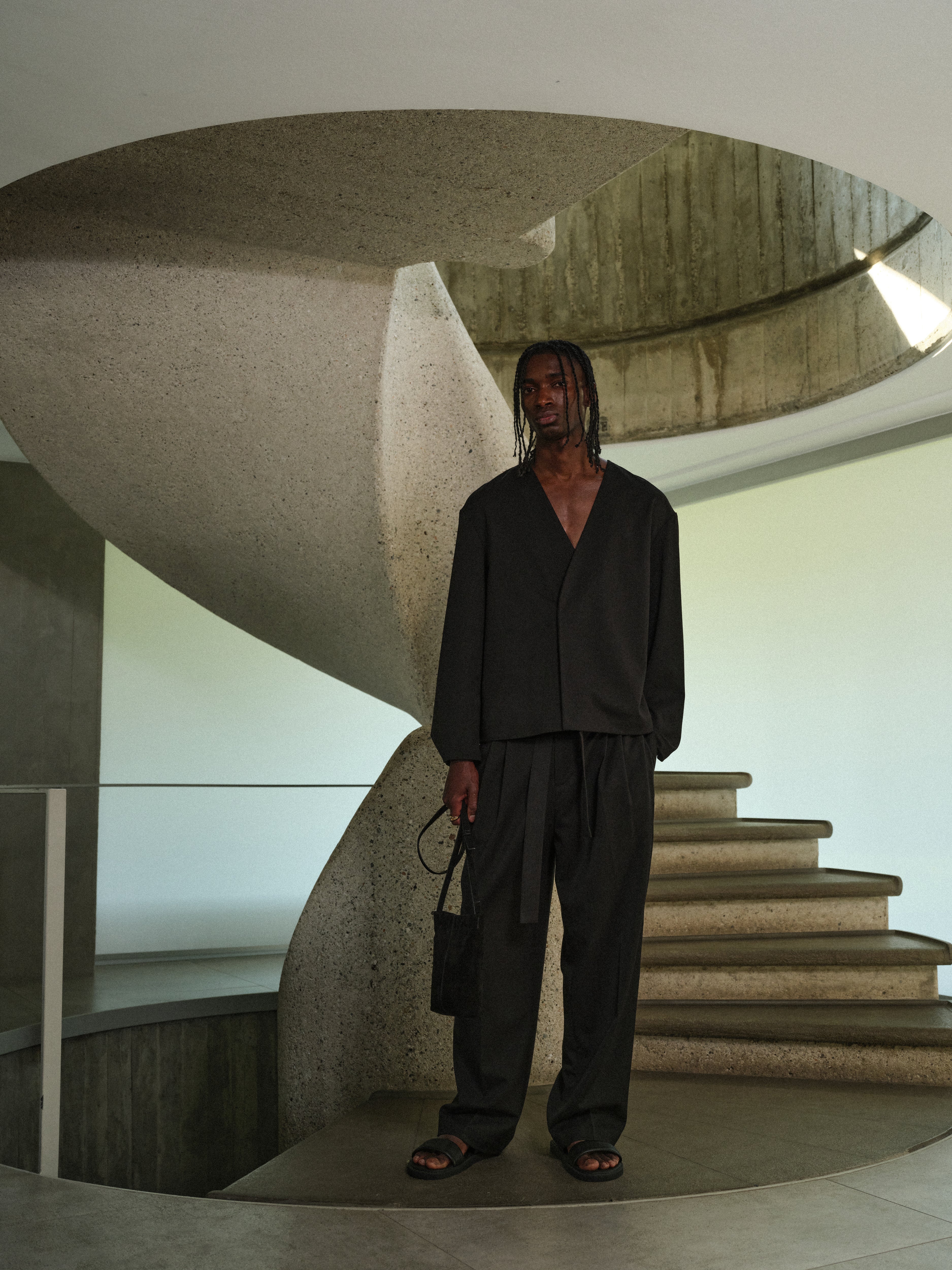 Man wearing black tailored trousers and a long sleeve tailored shirt, standing in a modern architectural setting with concrete walls and stairs.