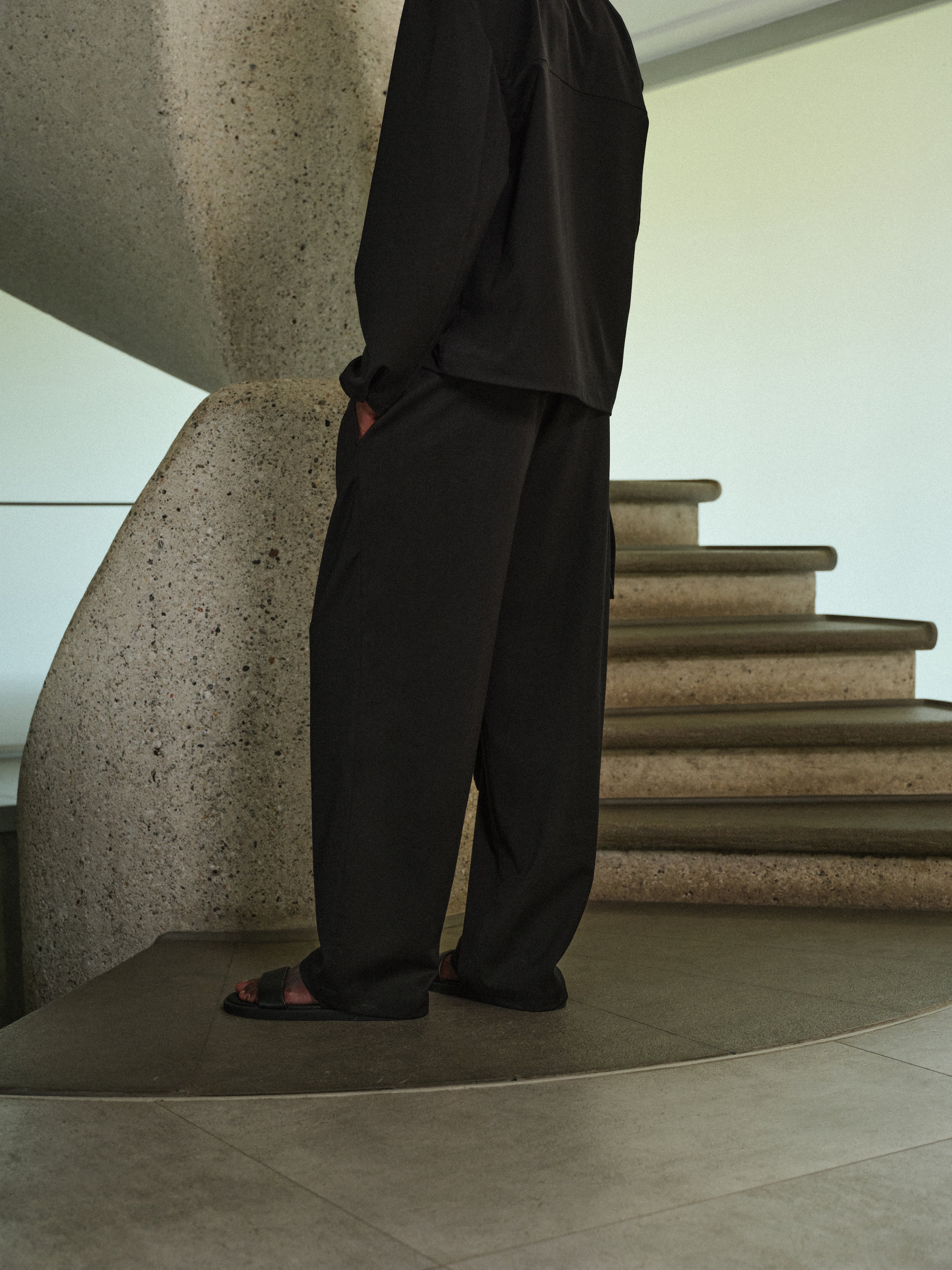 A man wearing black tailored trousers and a long sleeved tailored shirt, standing on a staircase with a modern architectural background