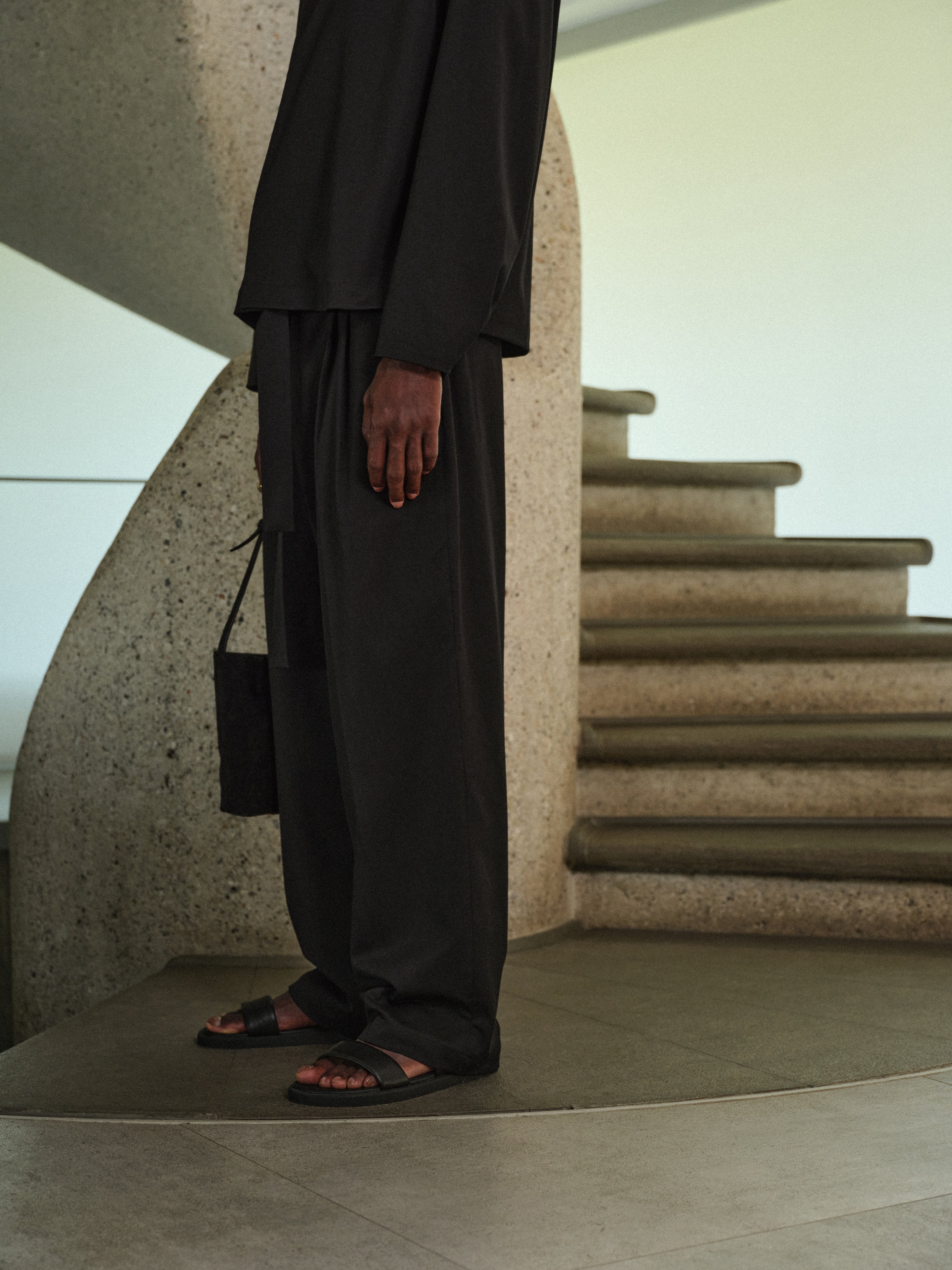 Man wearing black tailored trousers and sandals, standing by a spiral concrete staircase.