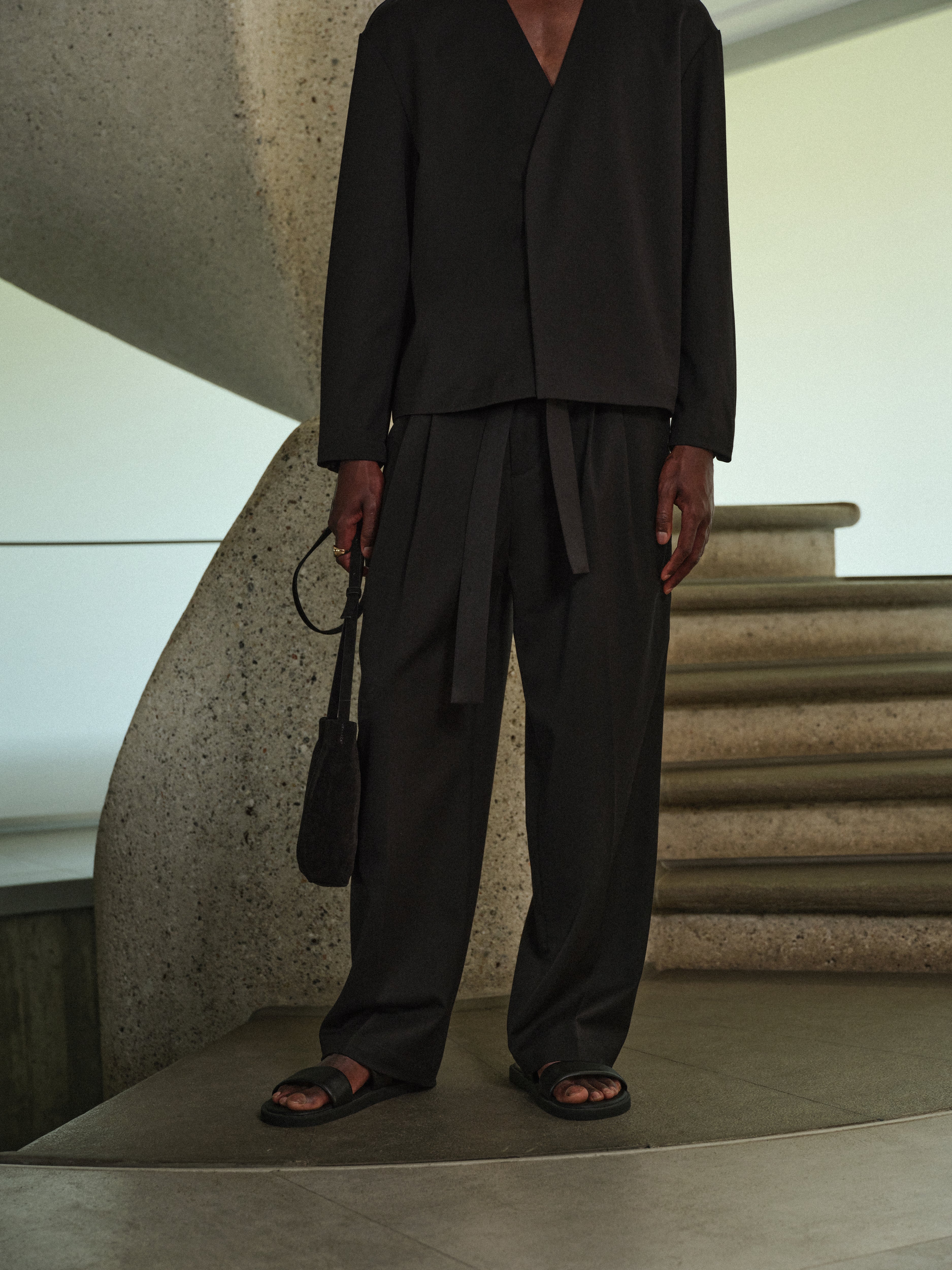 Man wearing black tailored trousers with a black long sleeved tailored shirt, standing on a staircase with concrete walls.