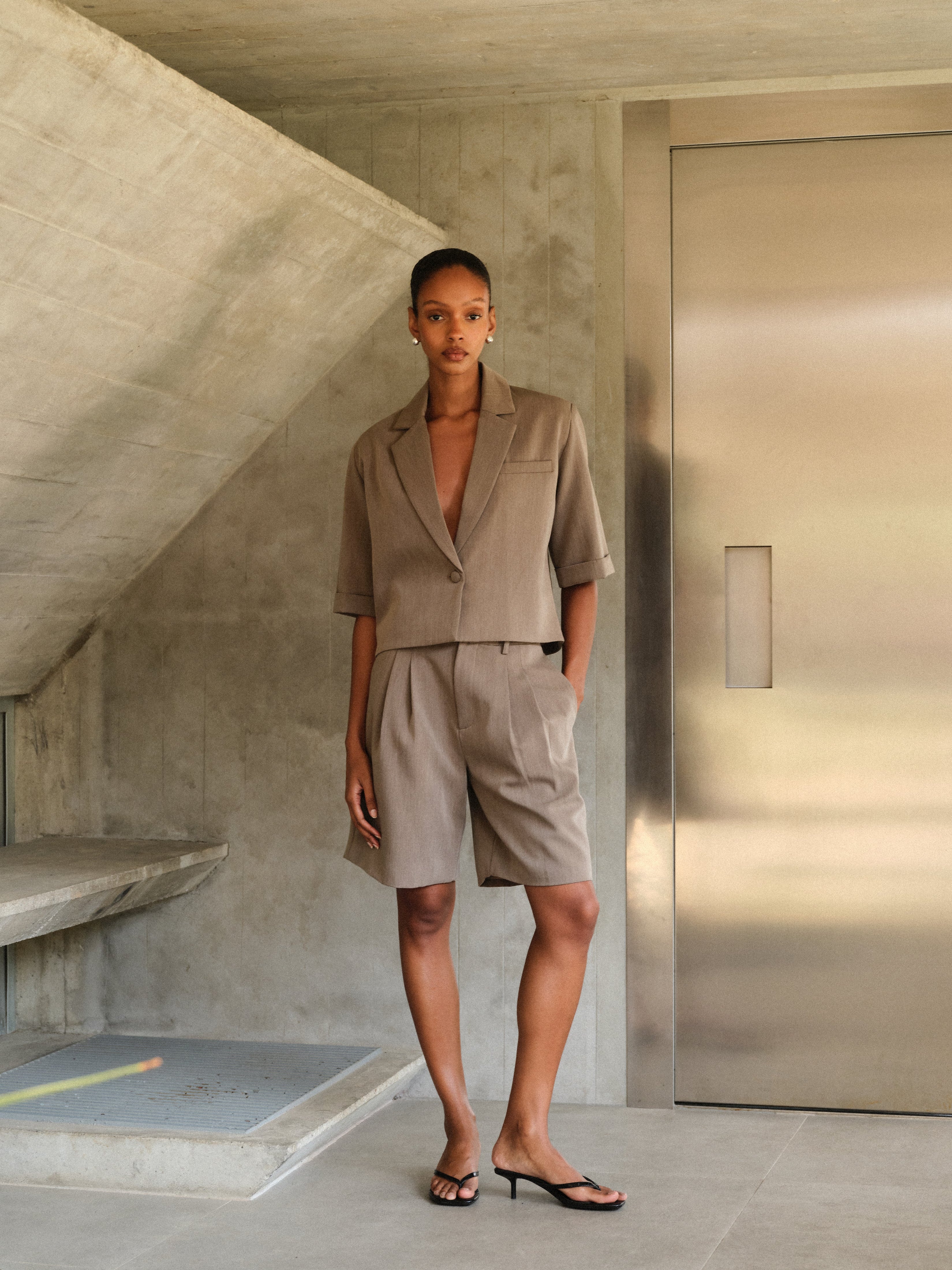 Woman in a beige tailored suit standing in a modern indoor setting with concrete walls and a metallic door.