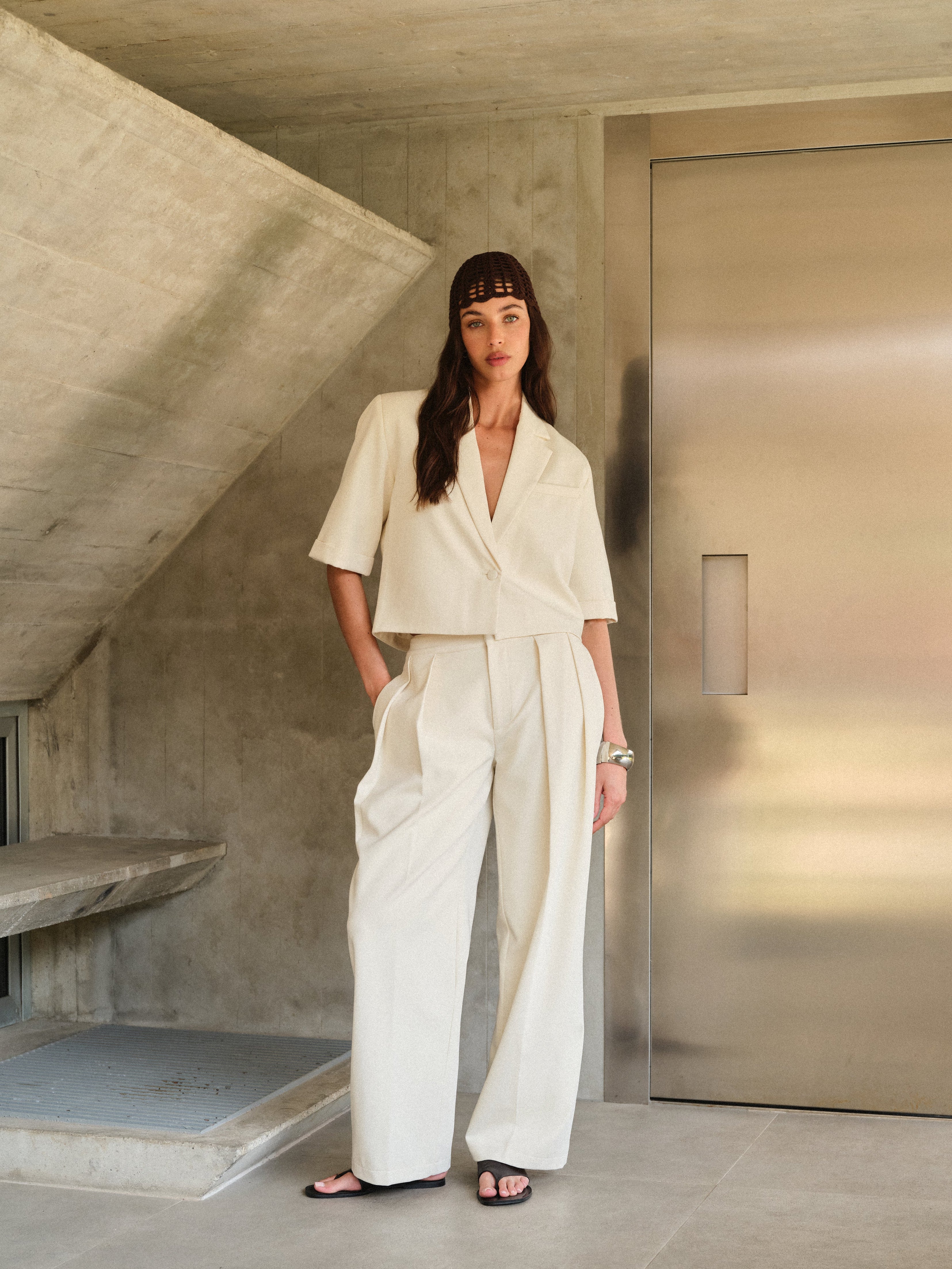 Woman wearing a white tailored blazer with brown a textured headband in an architectural setting with concrete walls and a metallic door.