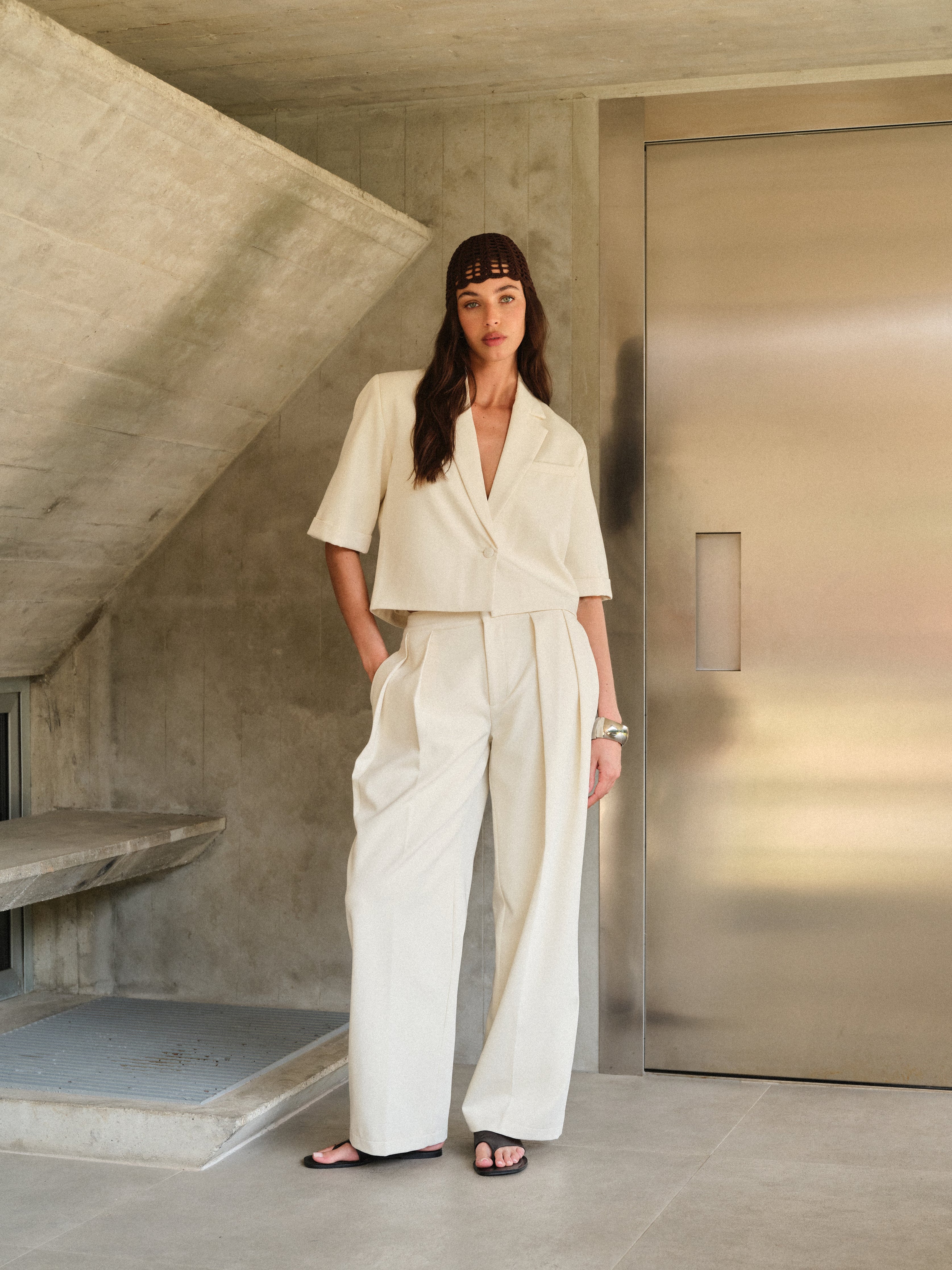 Woman wearing off white tailored trousers with matching blazer standing in a minimalistic room with concrete walls and a metallic door.