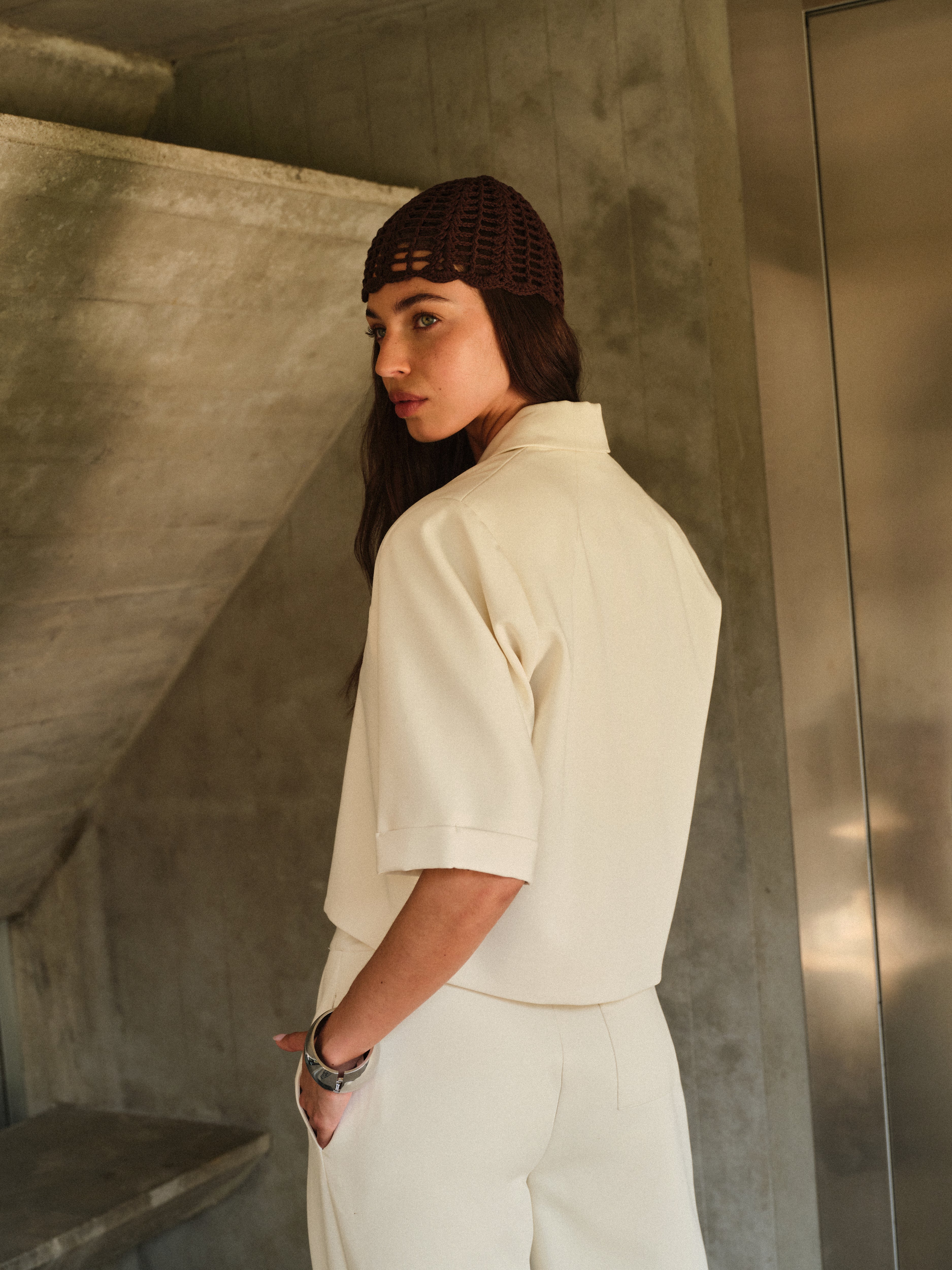 Woman wearing a white tailored blazer with brown a textured headband in an architectural setting with concrete walls and a metallic door.