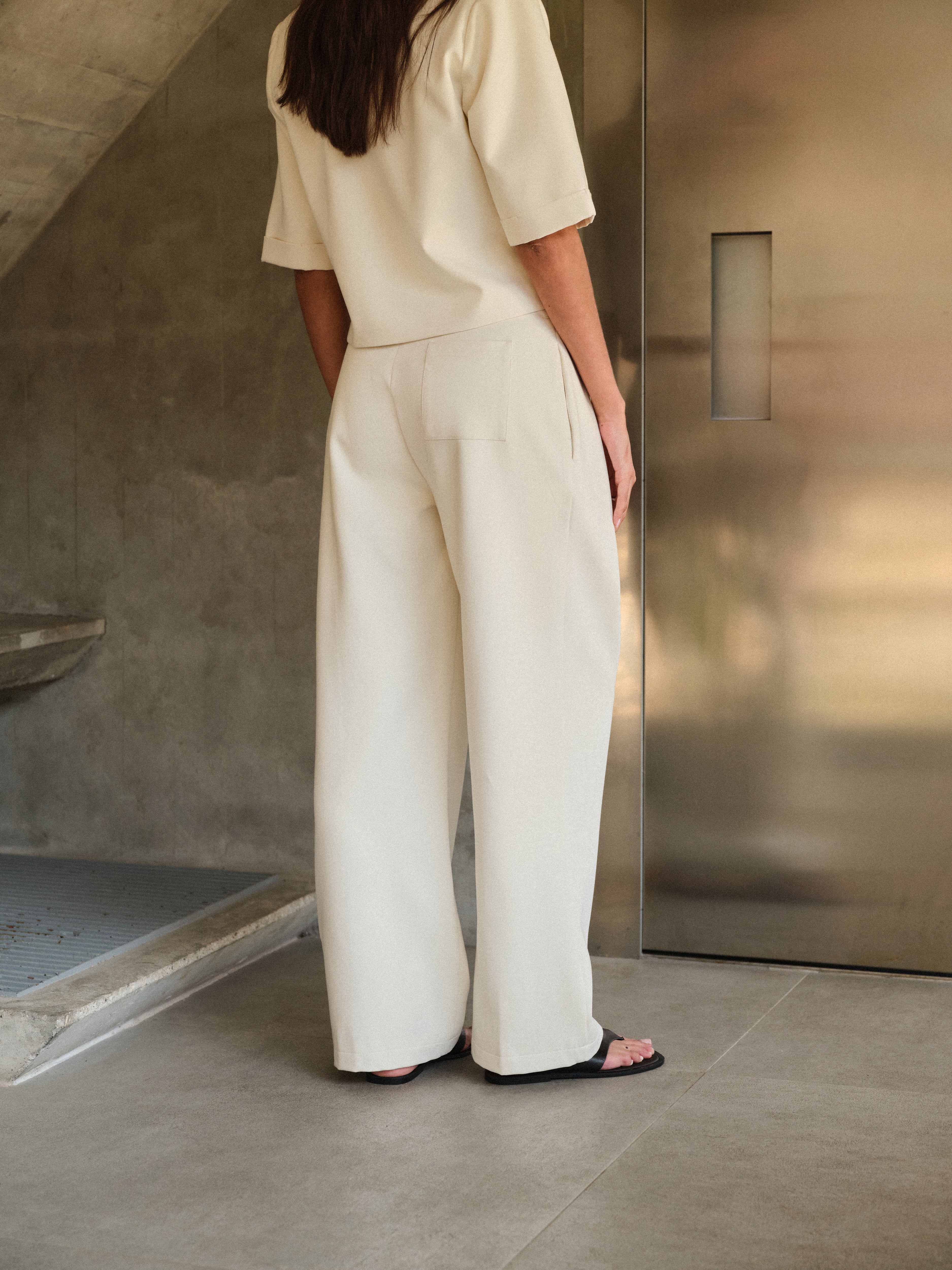 Woman wearing off white tailored trousers with matching blazer standing in a minimalistic room with concrete walls and a metallic door.