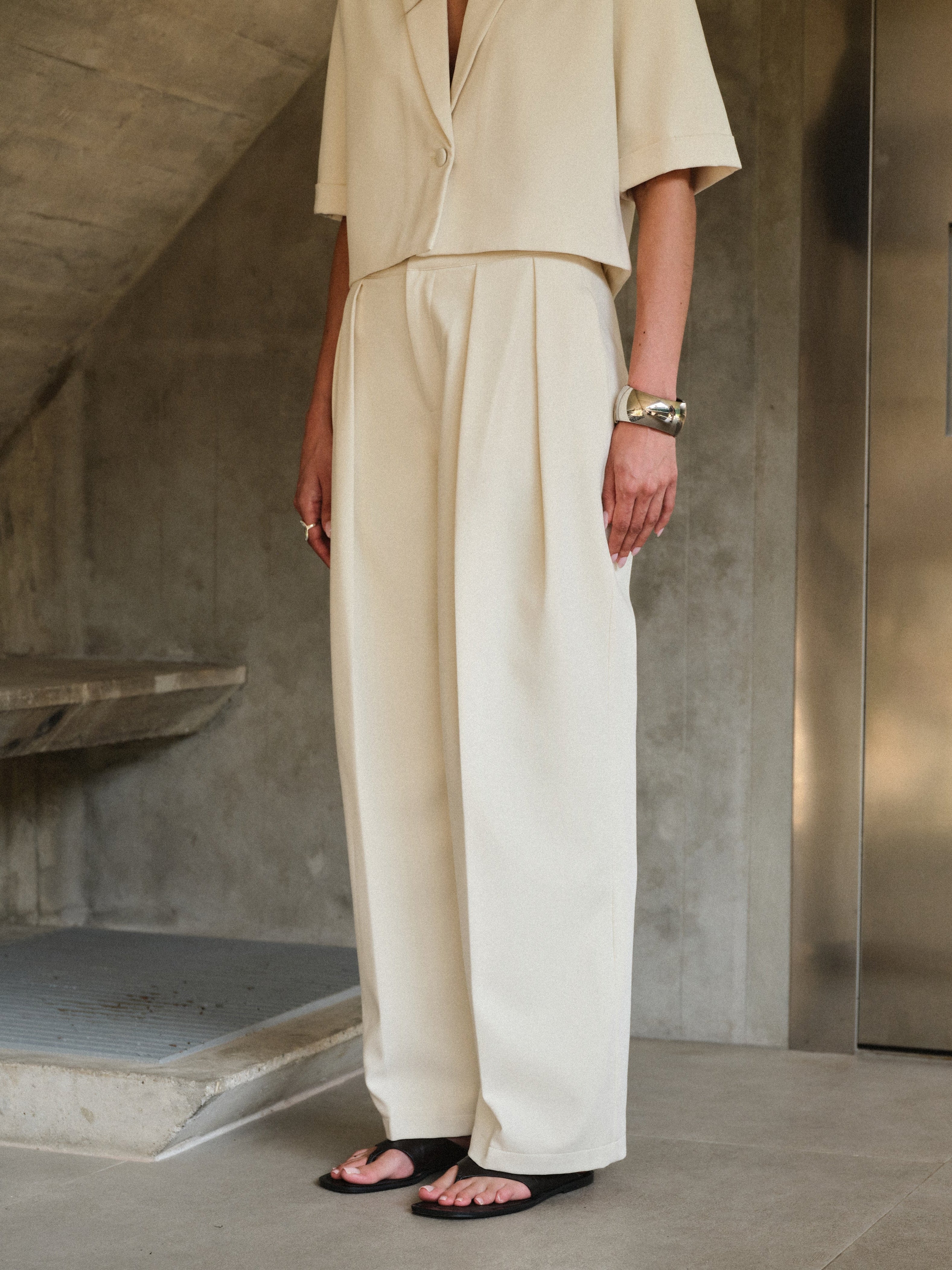 Woman wearing off white tailored trousers with matching blazer standing in a minimalistic room with concrete walls.