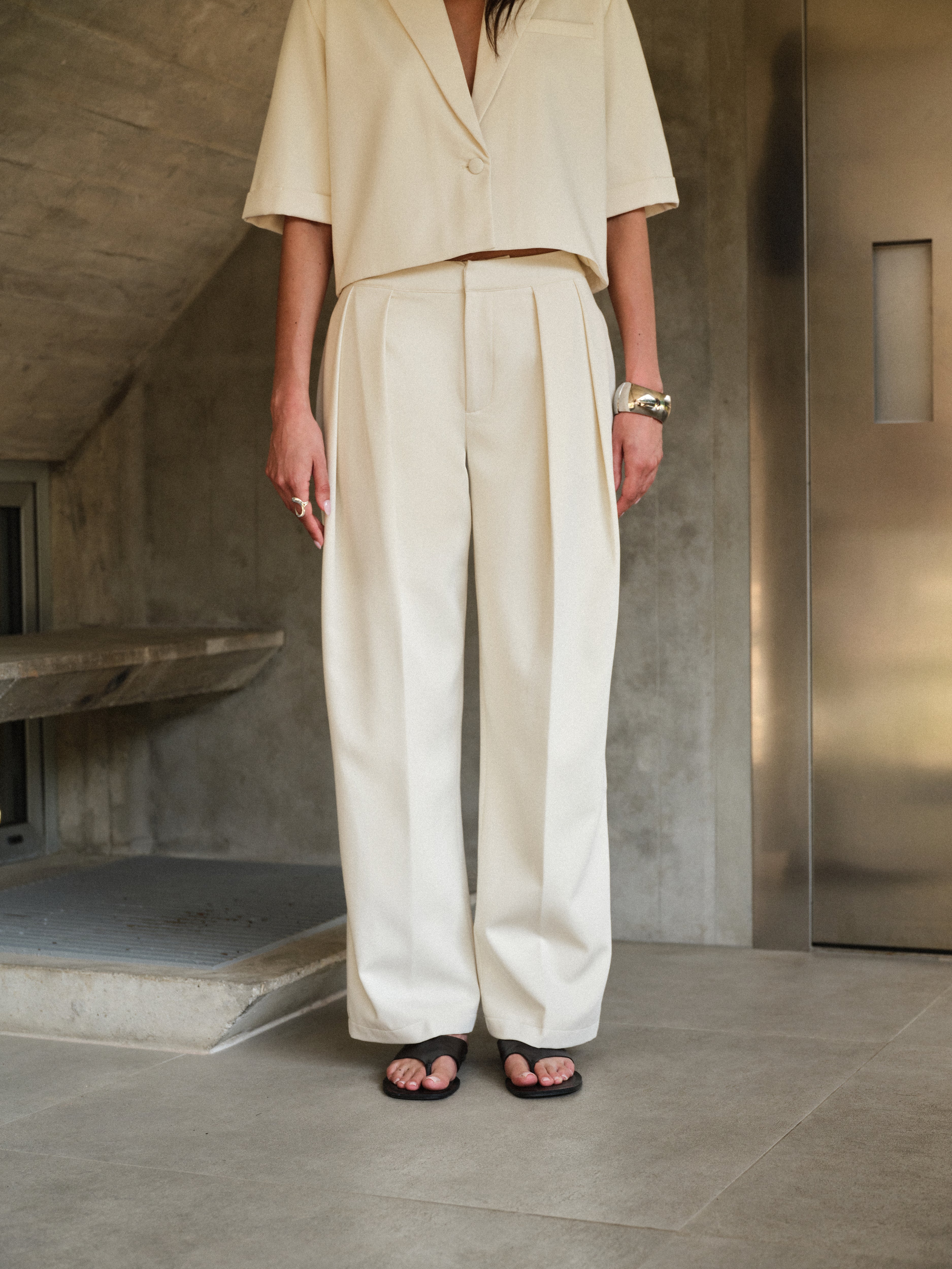 Woman wearing off white tailored trousers with matching blazer standing in a minimalistic room with concrete walls and a metallic door.