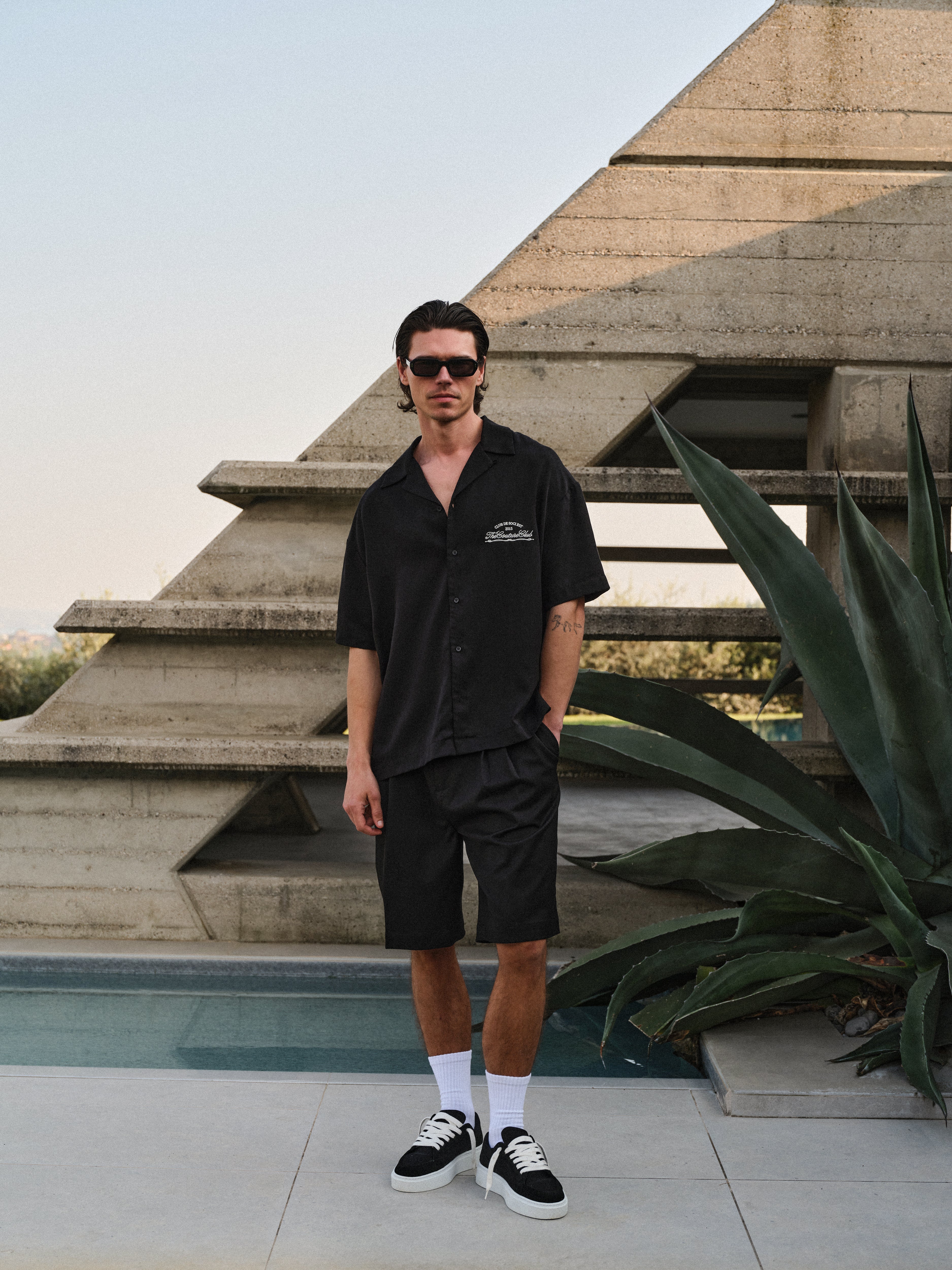 Man in black shirt and shorts standing in front of a modern architectural structure with a pool.