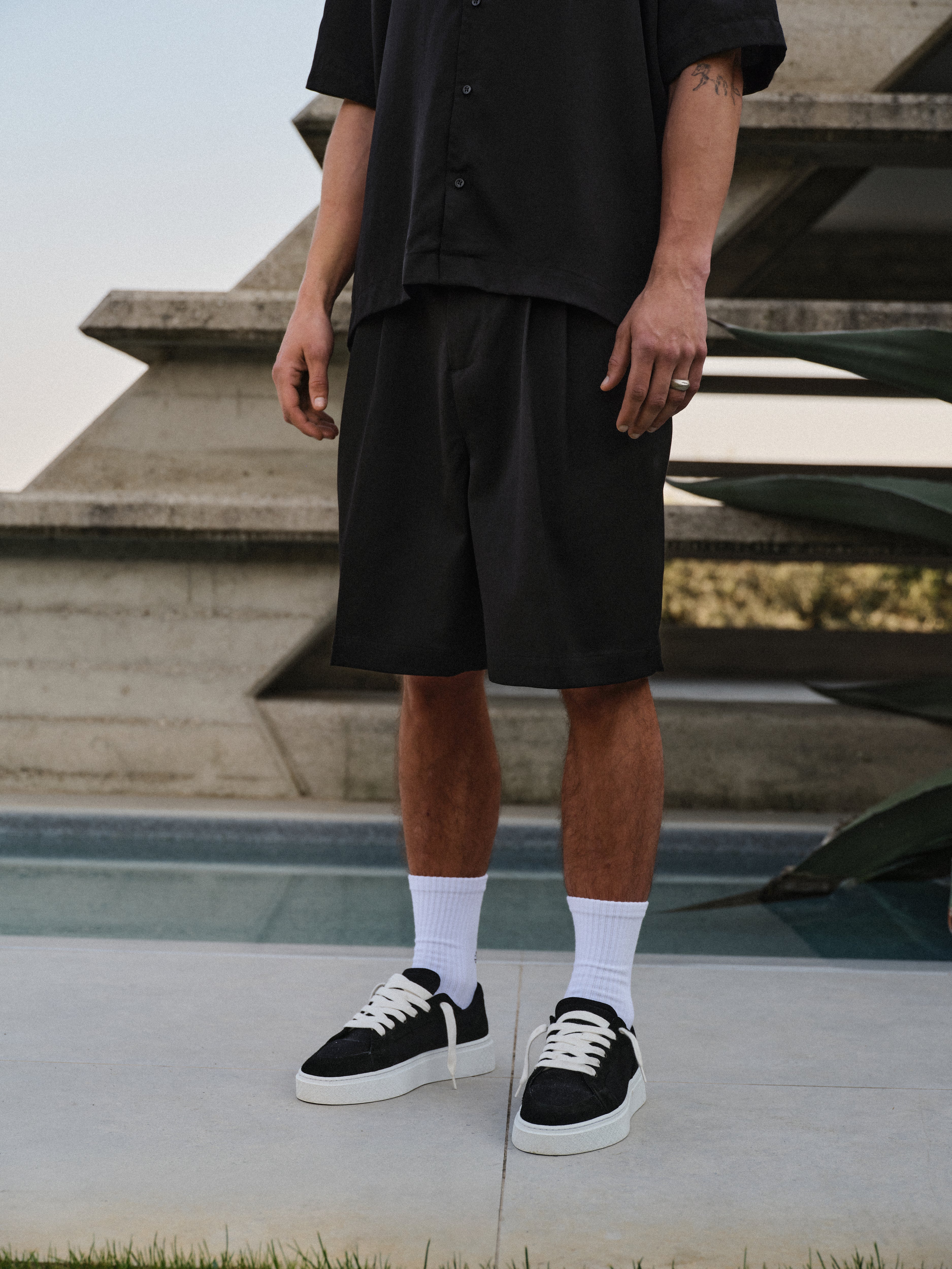 Man wearing a black outfit with shorts and sneakers standing on concrete floor.