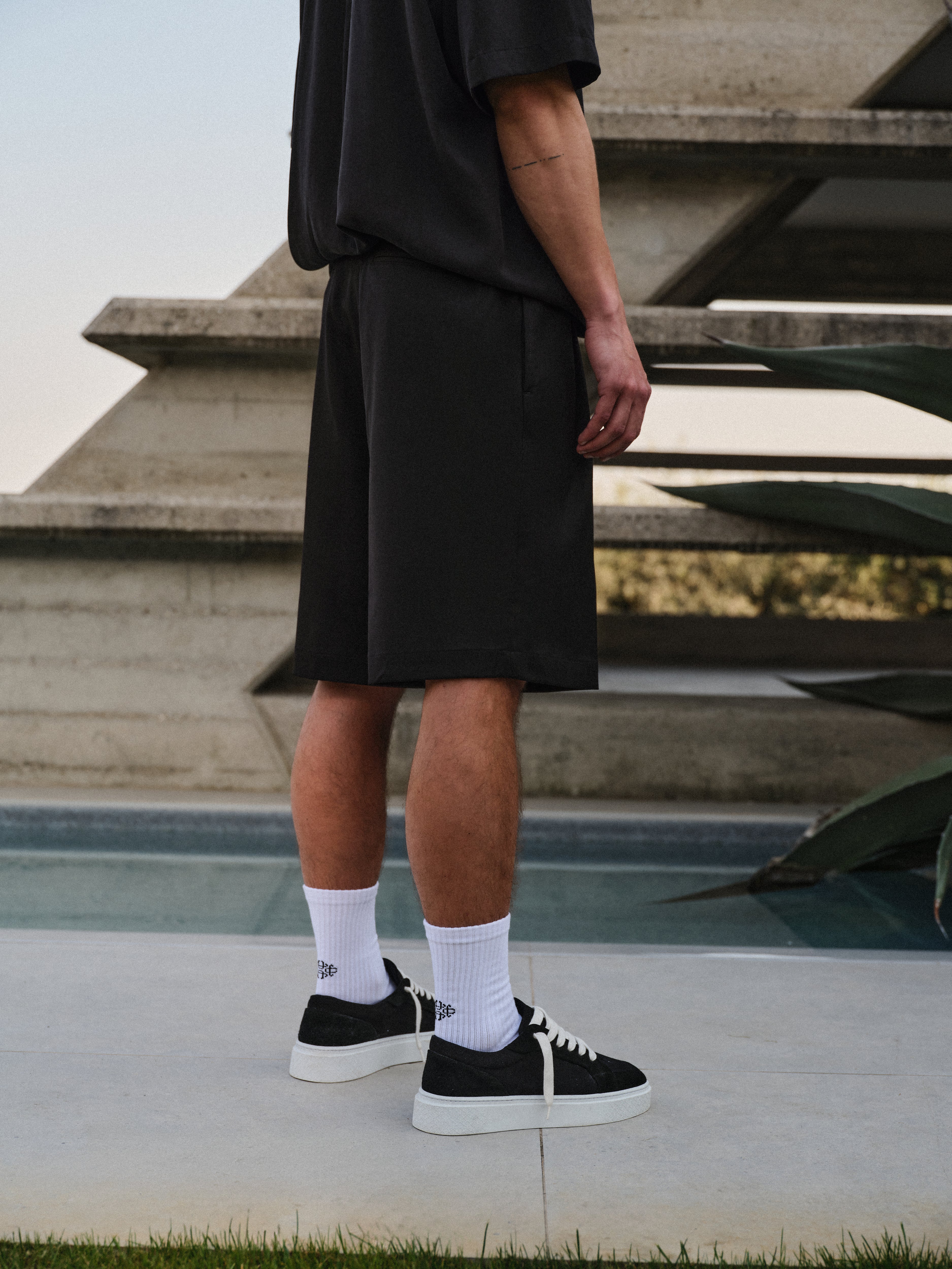 Man wearing black tailored shorts with a black shirt black, white socks and black sneakers standing on a concrete surface.