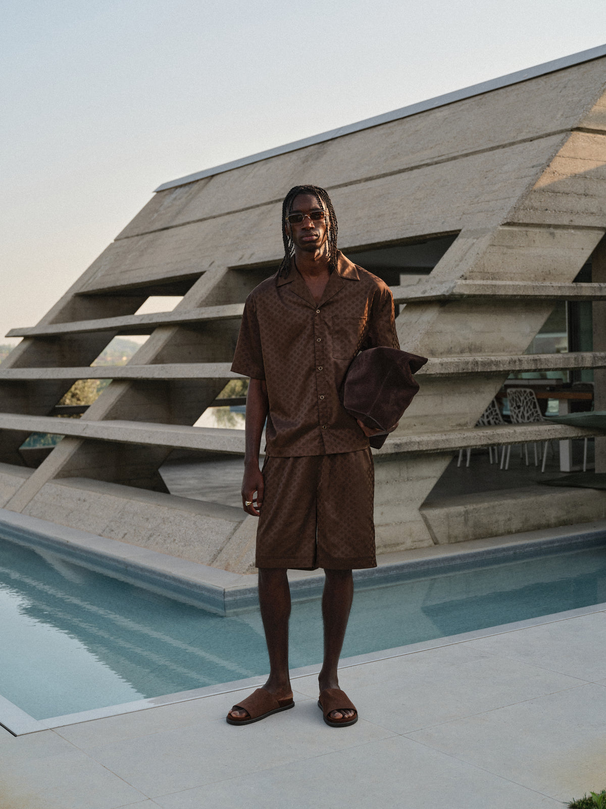 Man wearing brown satin shorts with matching brown sating shirt and sunglasses, standing by a pool with a geometric architectural background