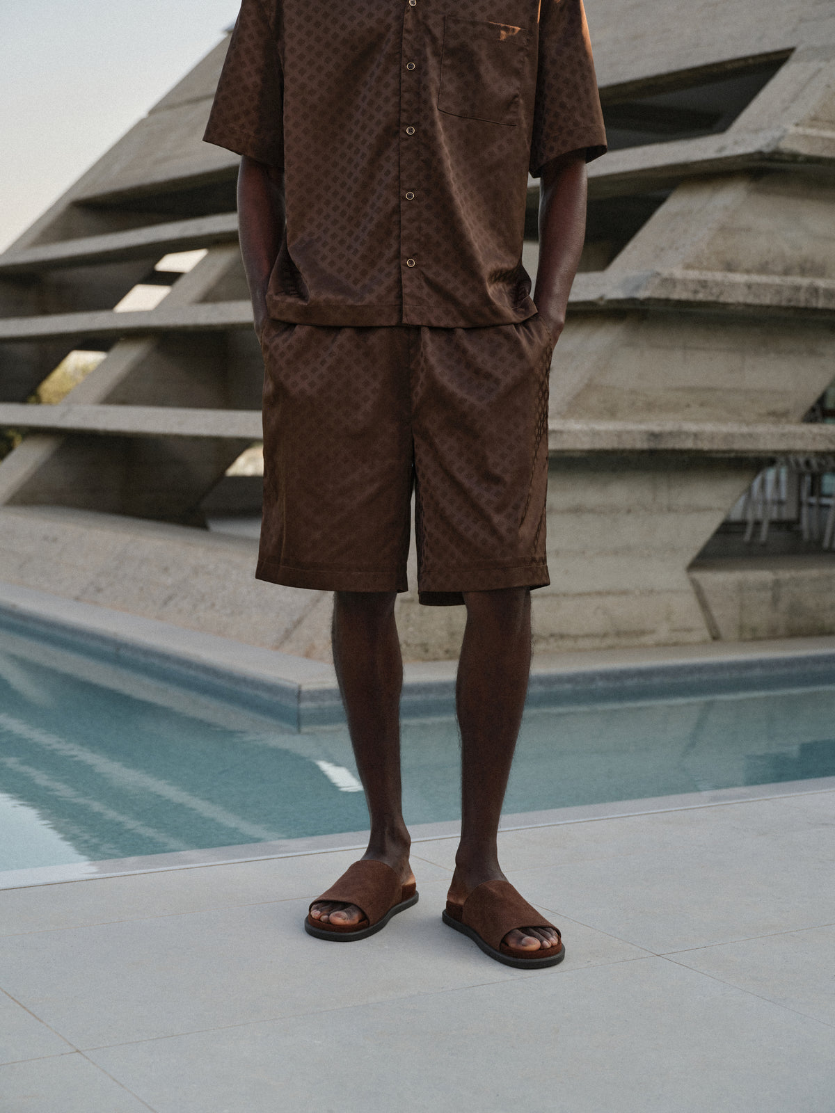 Man wearing brown satin shorts with matching brown sating shirt, standing by a pool with geometric architectural background