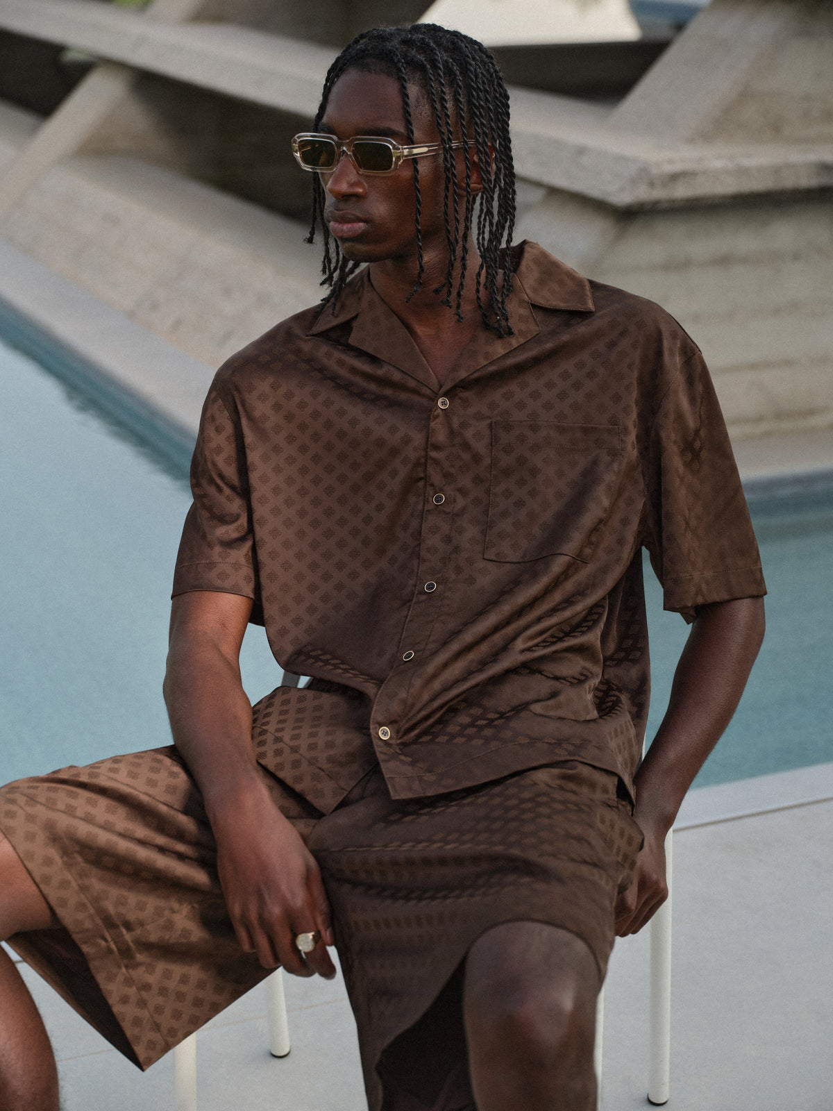 Man sat on a chair wearing brown satin shorts with a matching brown satin shirt, standing by a pool with geometric architectural background