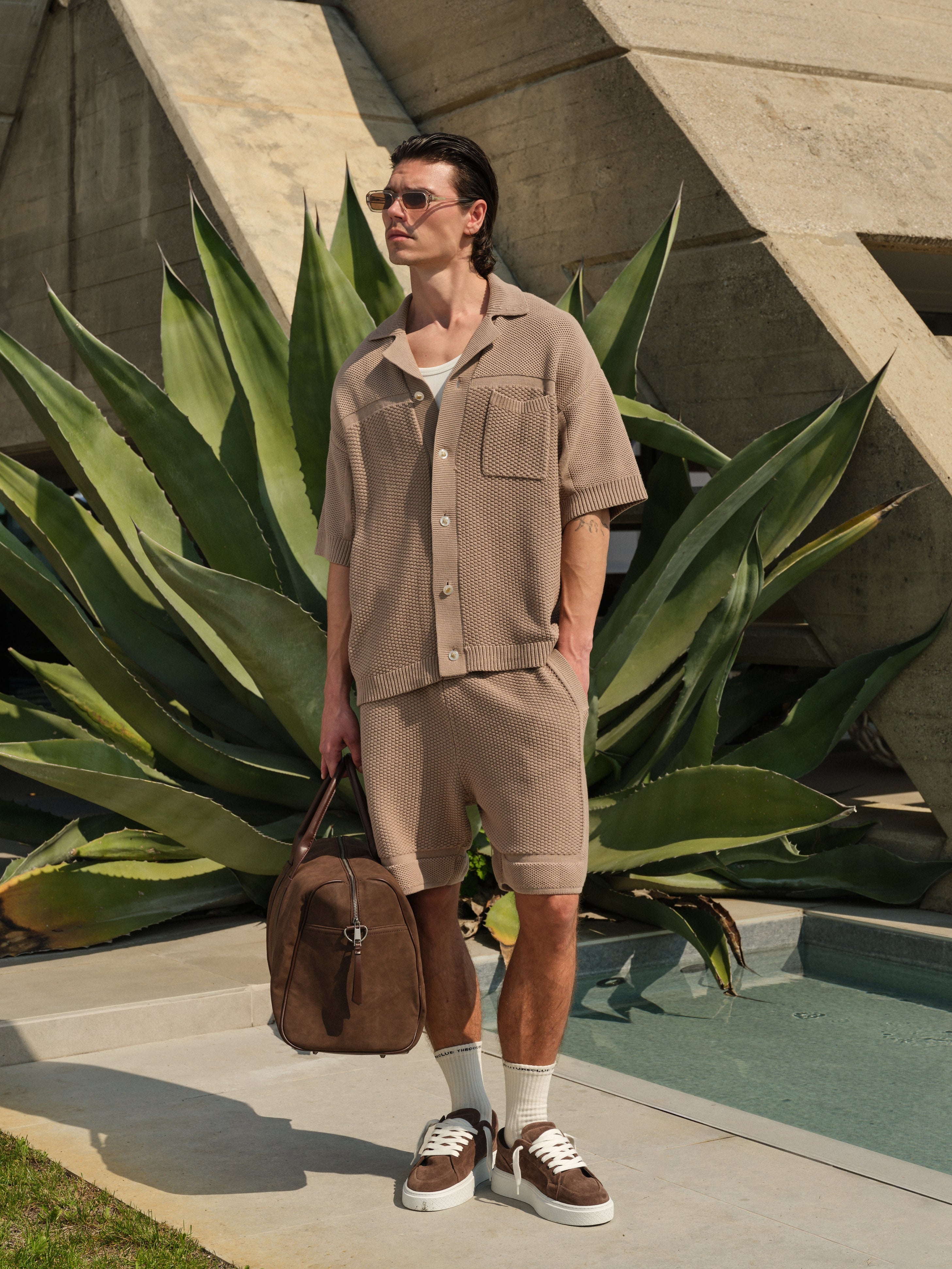 Man in beige outfit with brown backpack standing in front of a large plant