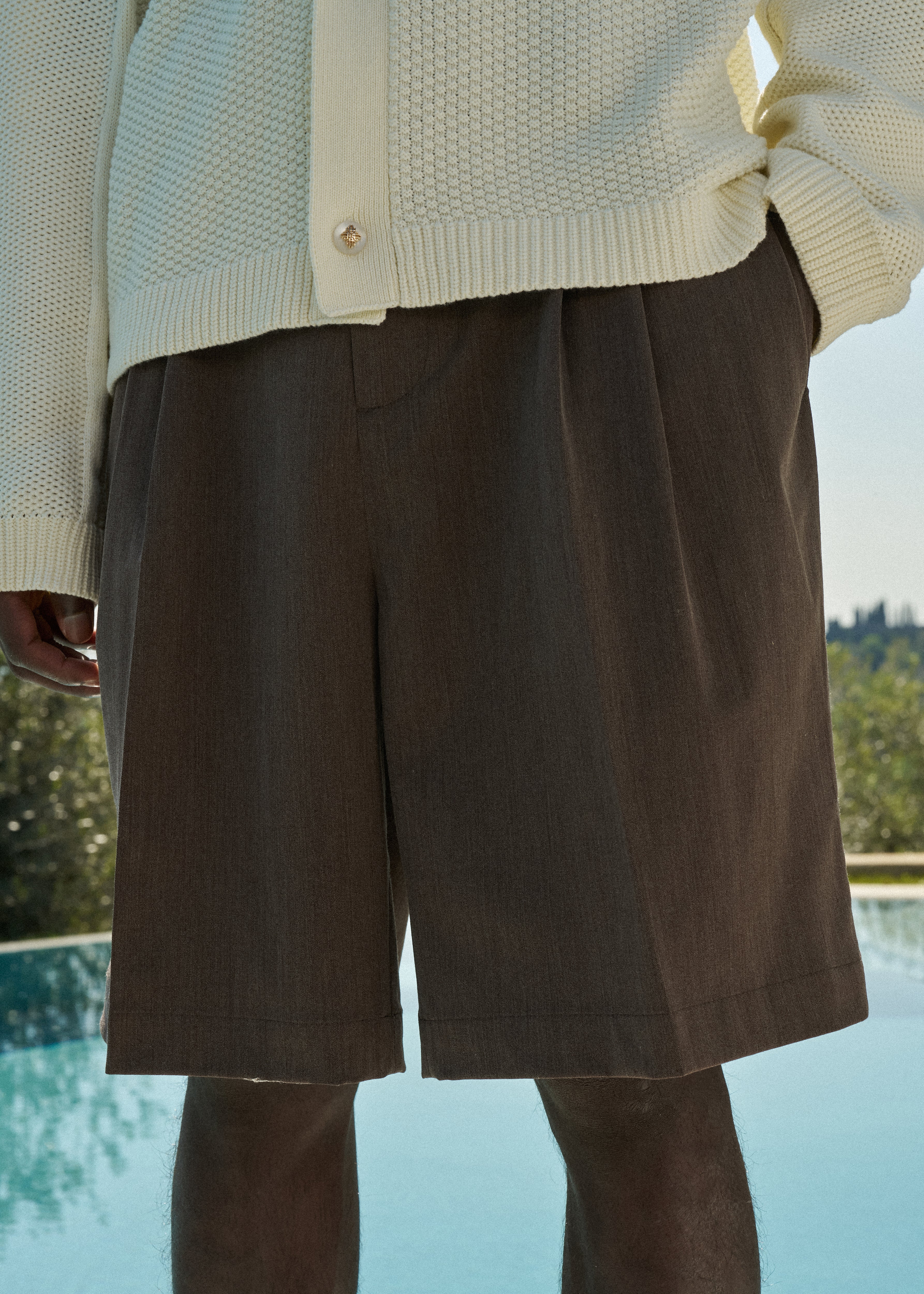 Close up of a man wearing brown tailored shorts and a white crochet knitted shirt by a poolside.