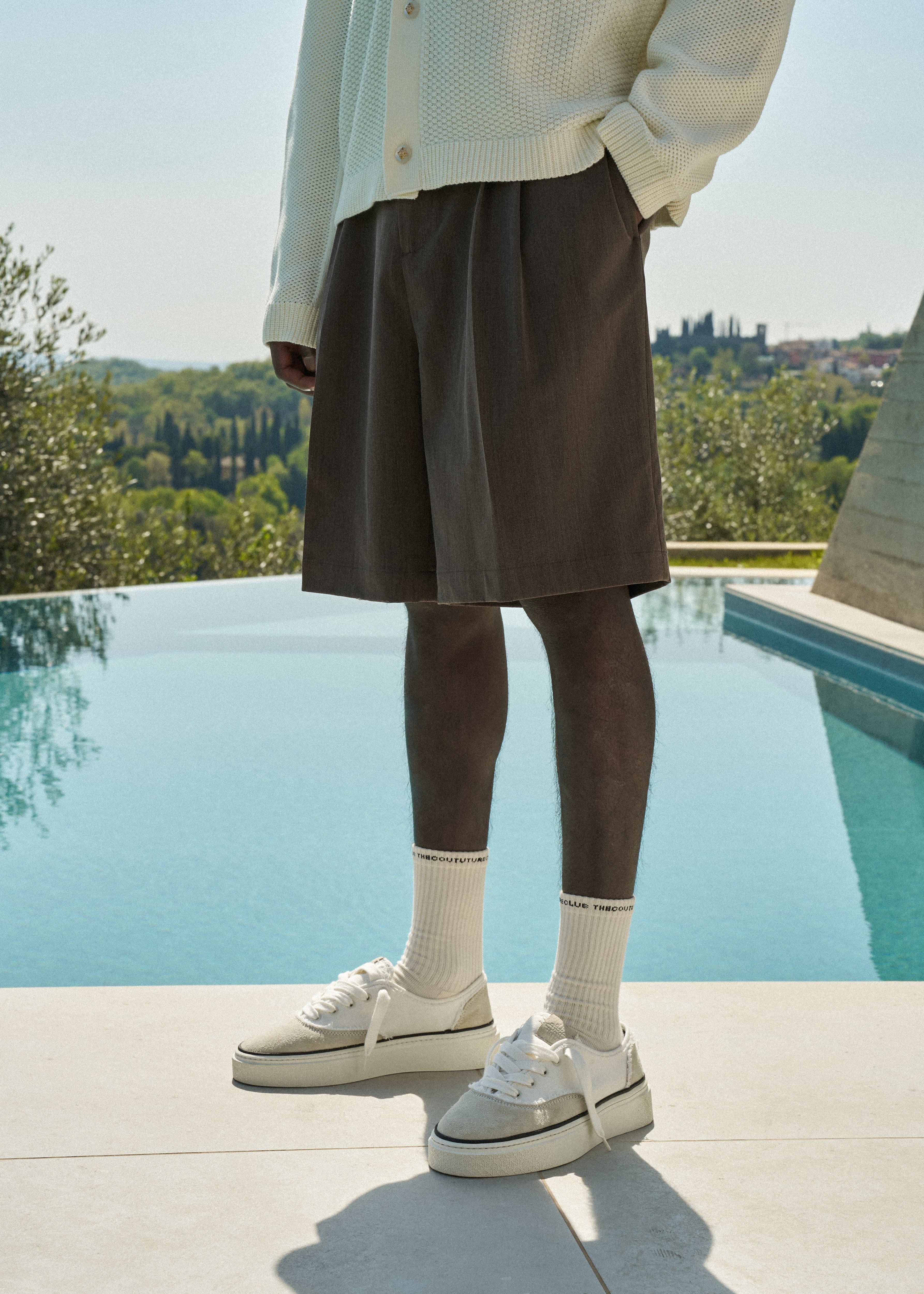 Man wearing brown tailored shorts, a white crochet knitted shirt and white sneakers by a pool with greenery in the background.