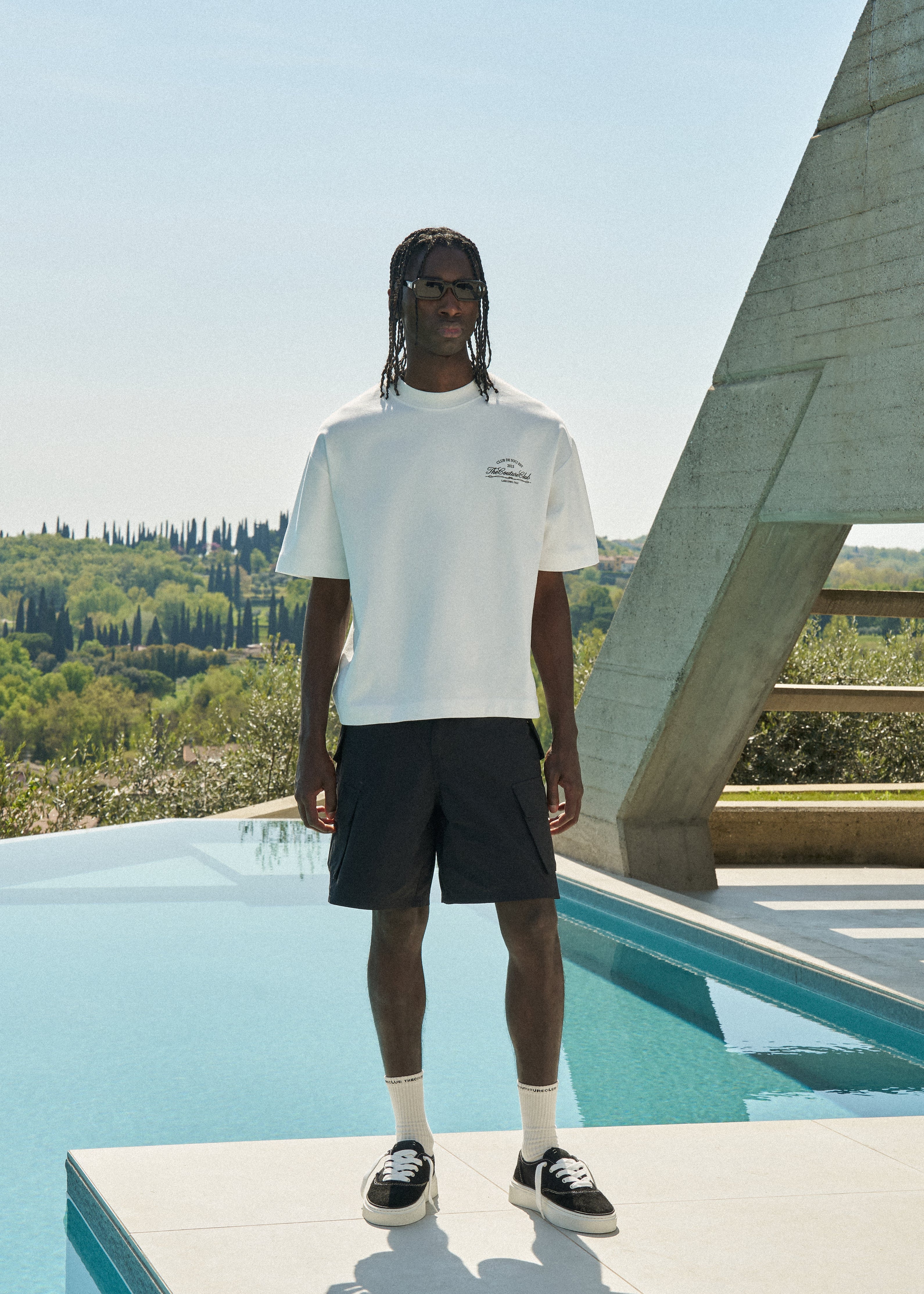 Man wearing a white t-shirt and black shorts standing by a pool with a scenic background