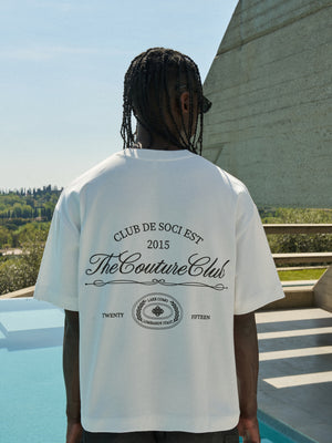 Person wearing a white t-shirt with 'The Couture Club' design, standing by a pool with a scenic background.