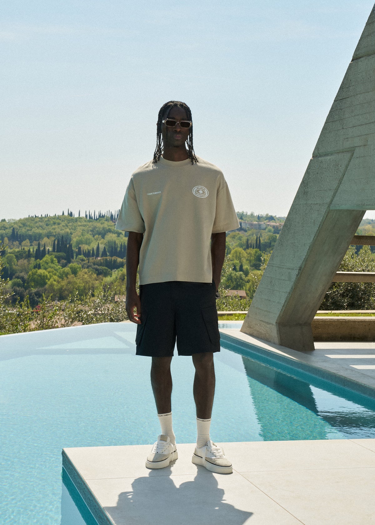 Man standing by a pool in a beige t-shirt and sunglasses with a scenic background
