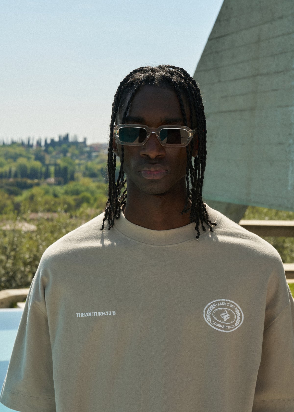 Man wearing sunglasses and a beige t-shirt with logos, standing outdoors.