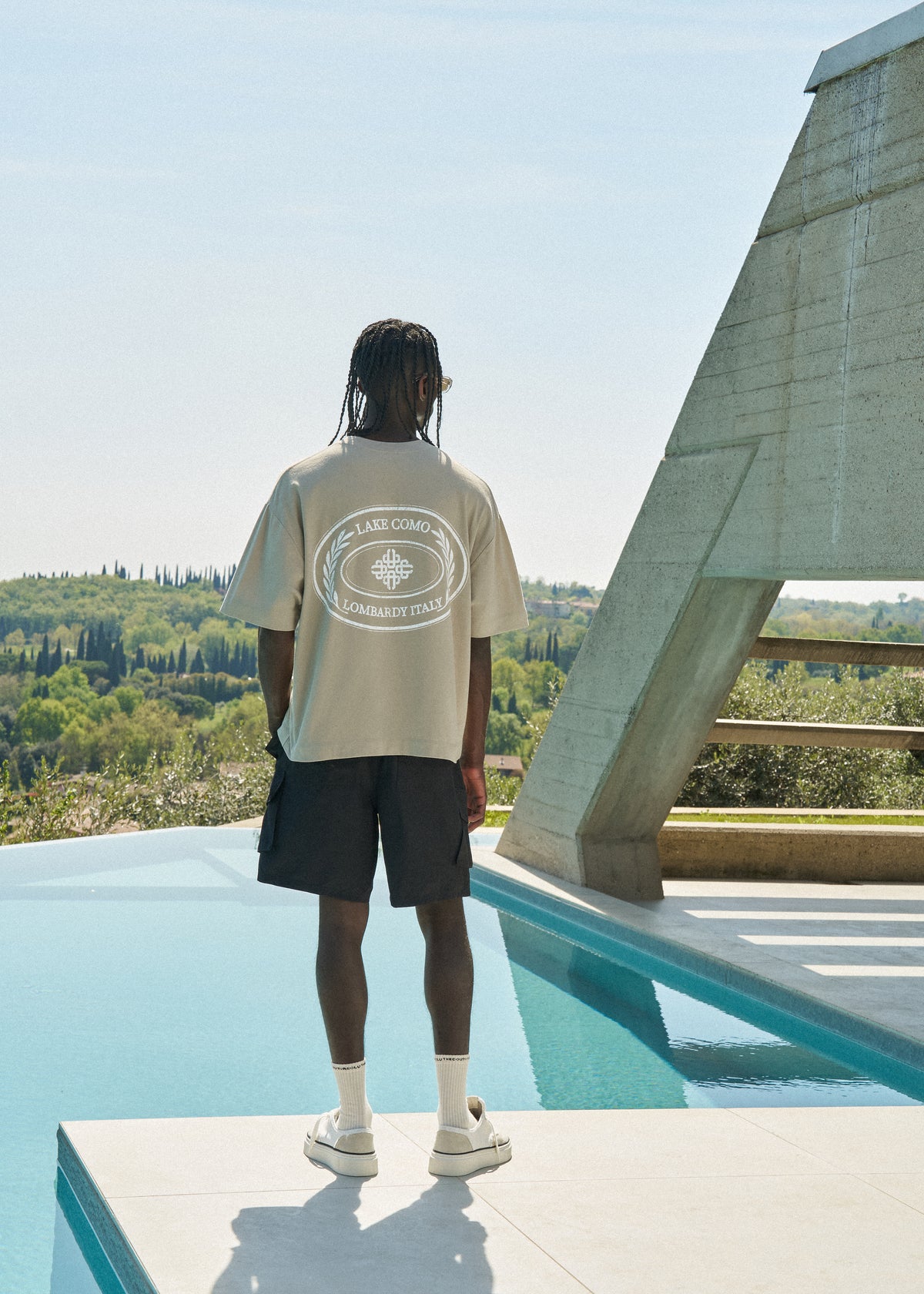 Man wearing a beige t-shirt and black shorts standing on a rooftop pool deck with a scenic view