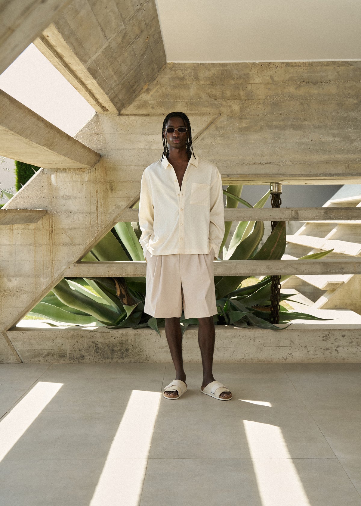 Man wearing beige tailored shorts, a white satin long-sleeved shirt in a modern architectural setting with plants.