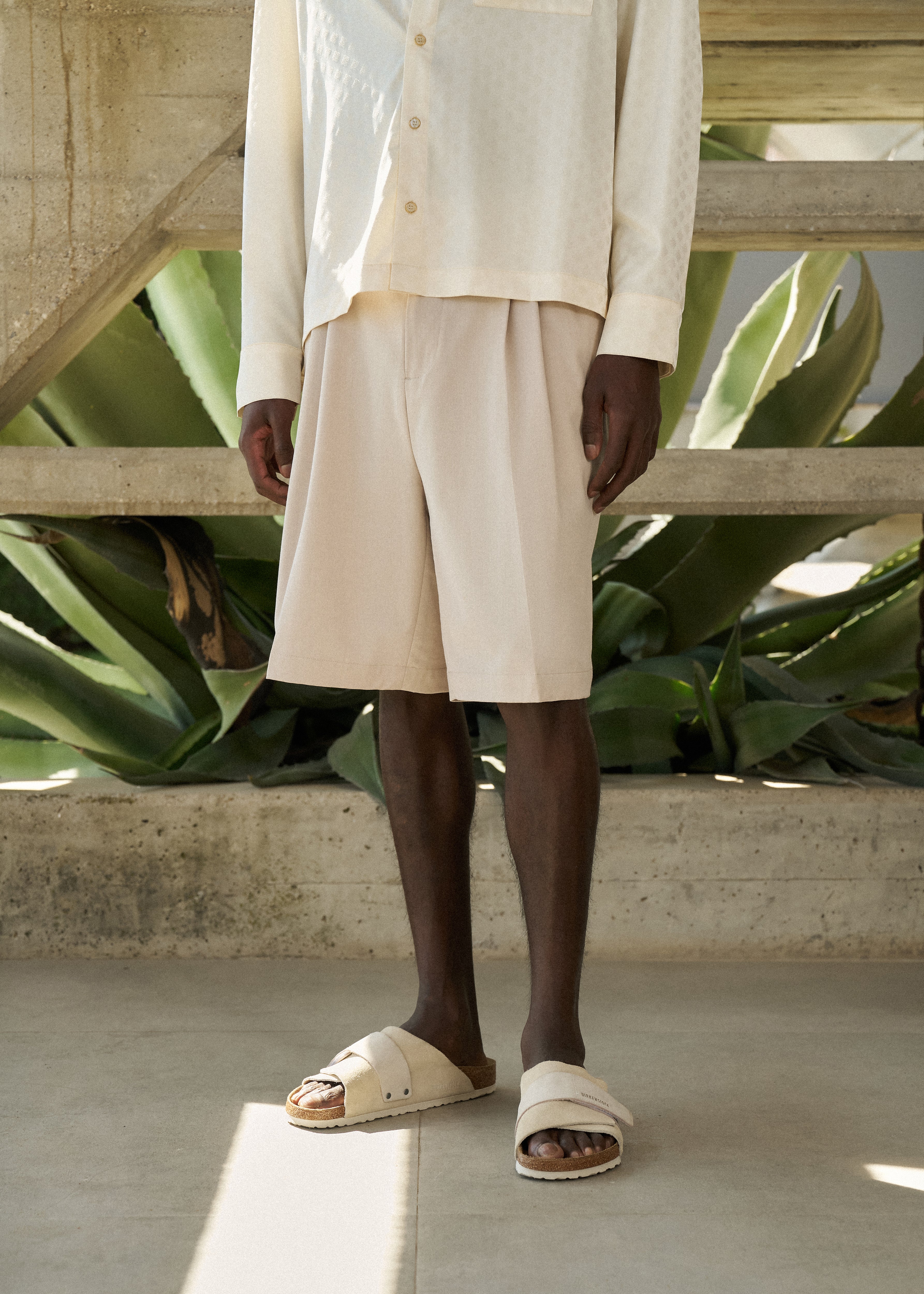 Man wearing beige tailored shorts, with a satin long-sleeved satin shirt outfit with sandals standing in front of green plants.