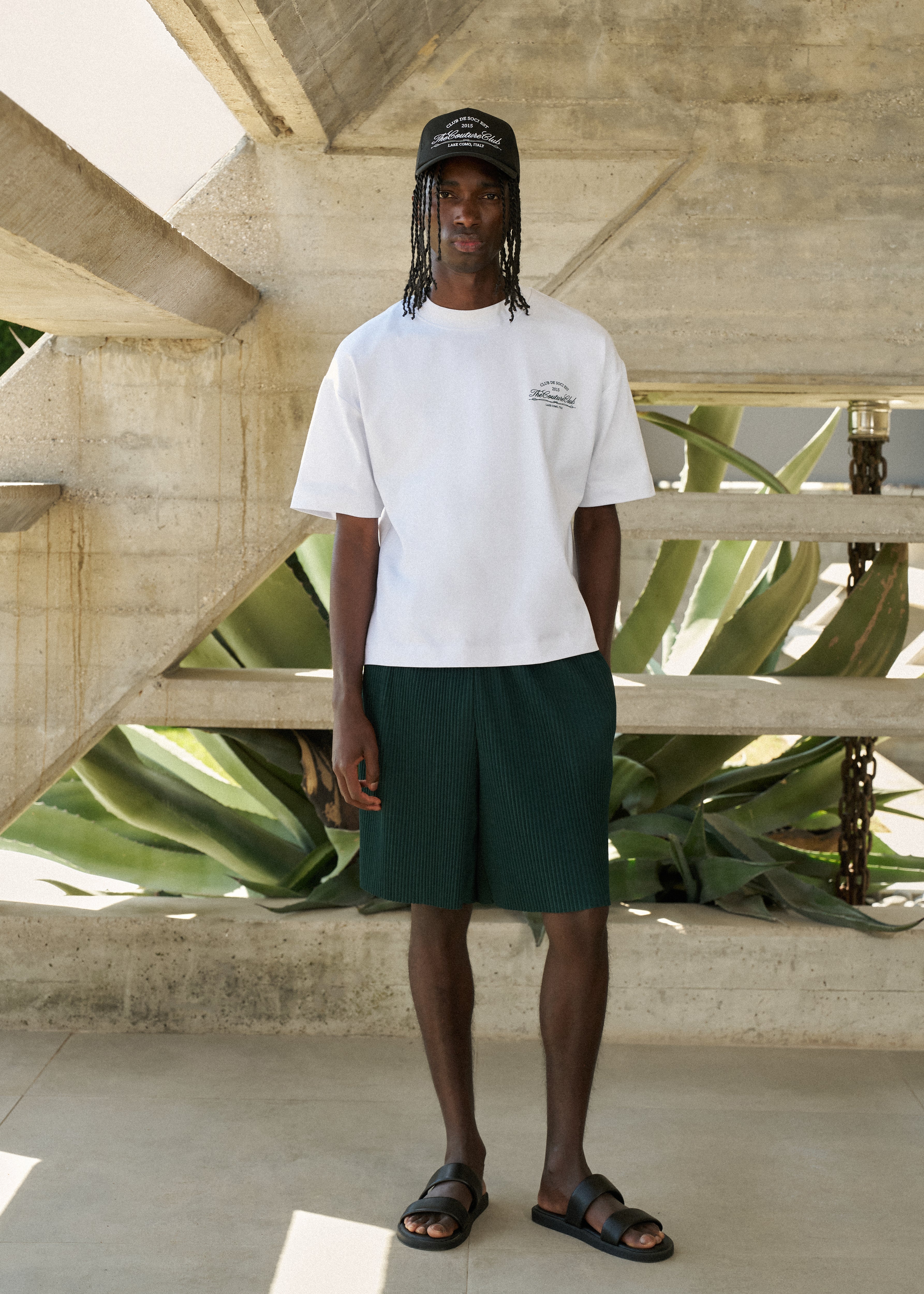 Person wearing a white t-shirt and green shorts standing in front of a concrete structure with plants.