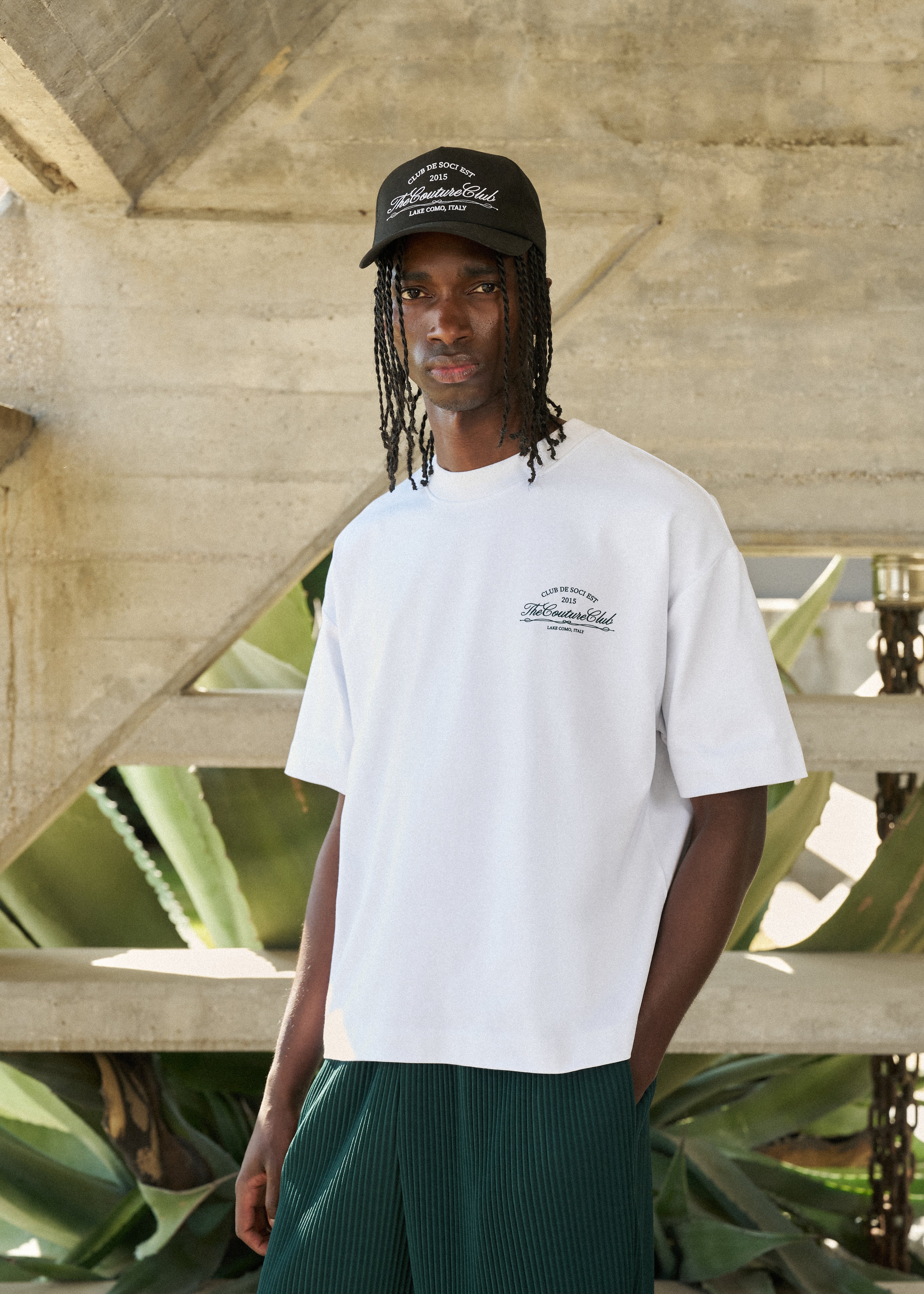 Person wearing a white t-shirt with a logo and a black cap, standing in front of a concrete wall and plants.
