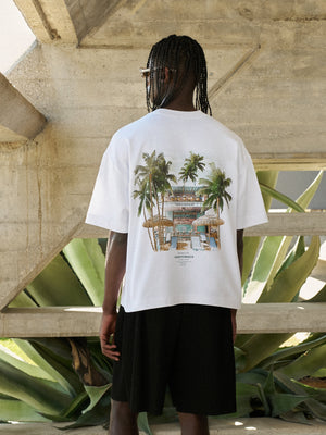 Person wearing a white t-shirt with a tropical design, standing in front of a concrete structure with plants.