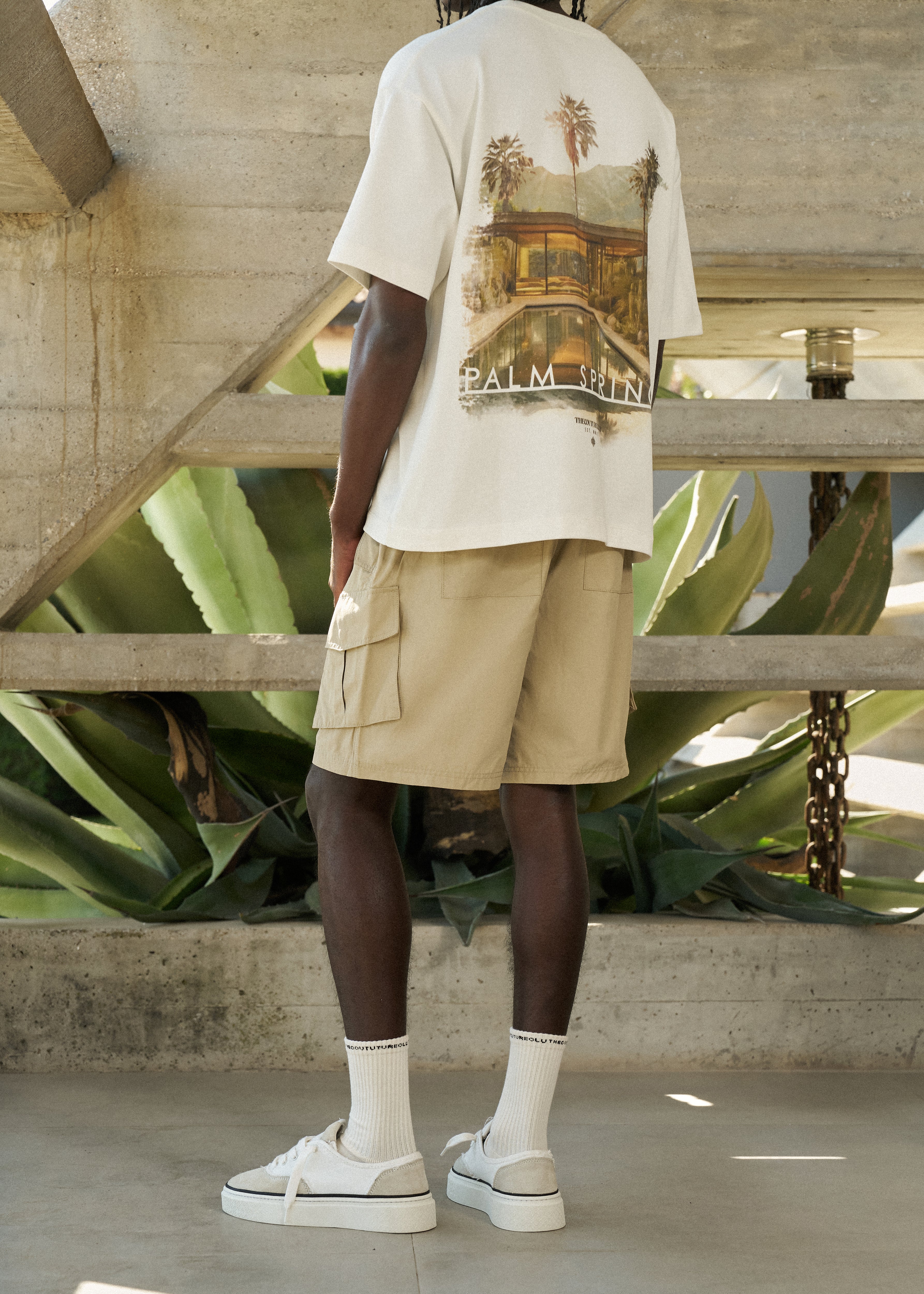 Man wearing a beige t-shirt with a graphic design and beige cargo shorts, standing in an outdoor setting.