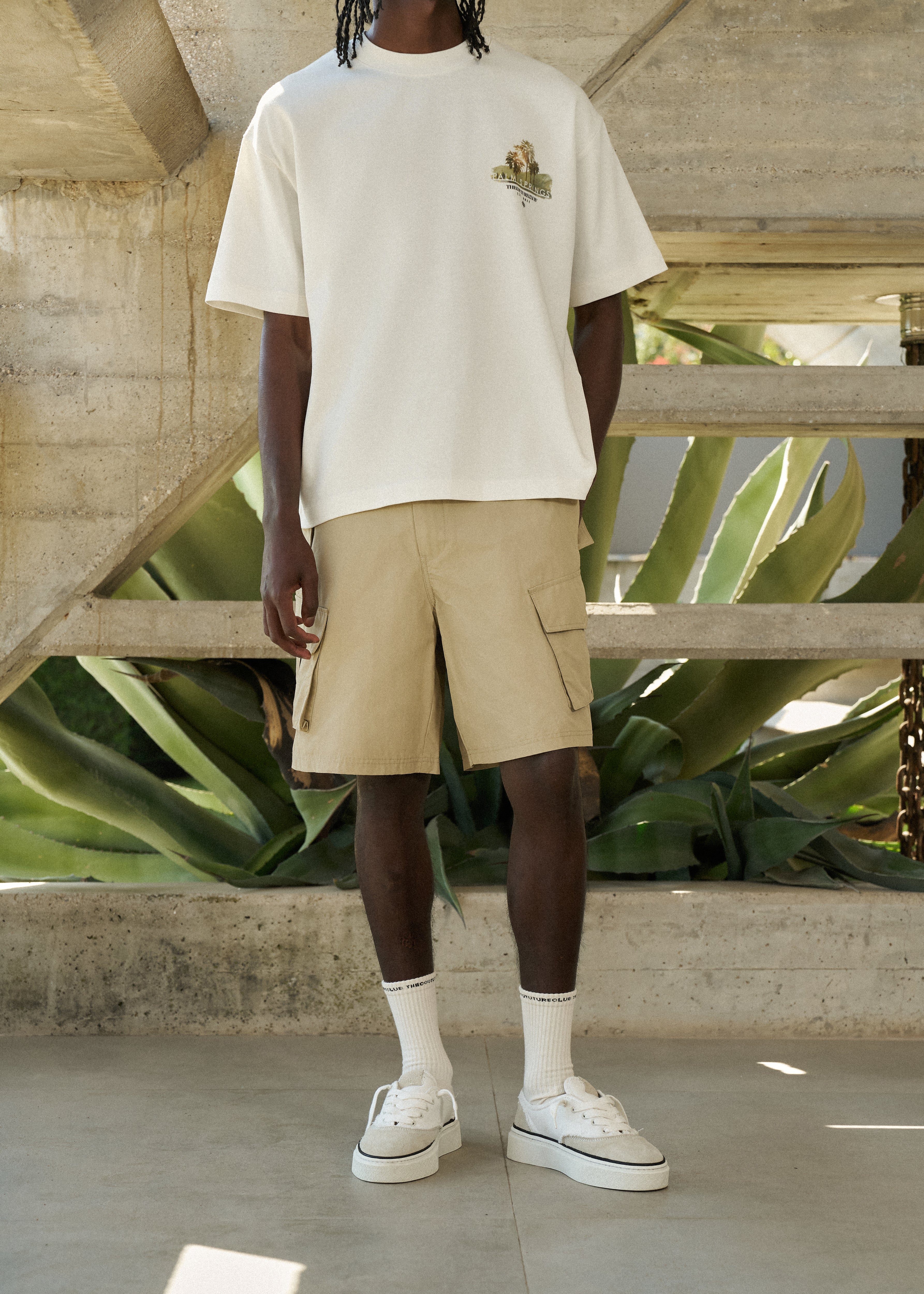 Man wearing a white t-shirt, beige cargo shorts, and white sneakers in an outdoor setting with plants and concrete.