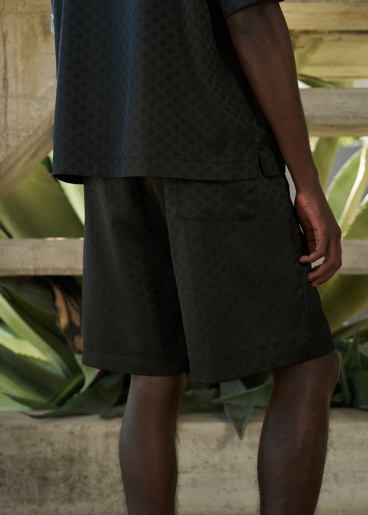 Man wearing black satin shorts, with matching satin shirt standing in front of large green leaves.