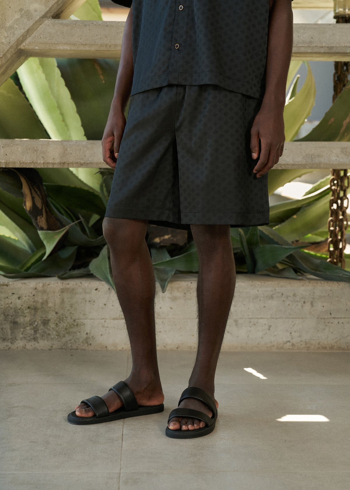 Man wearing black satin shorts, with matching satin shirt with sandals standing in front of large green leaves.