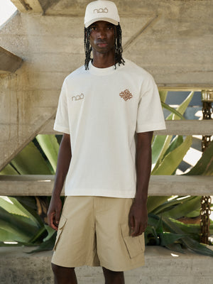 Man wearing a white t-shirt and beige shorts with branding, standing against a concrete structure and plant background.