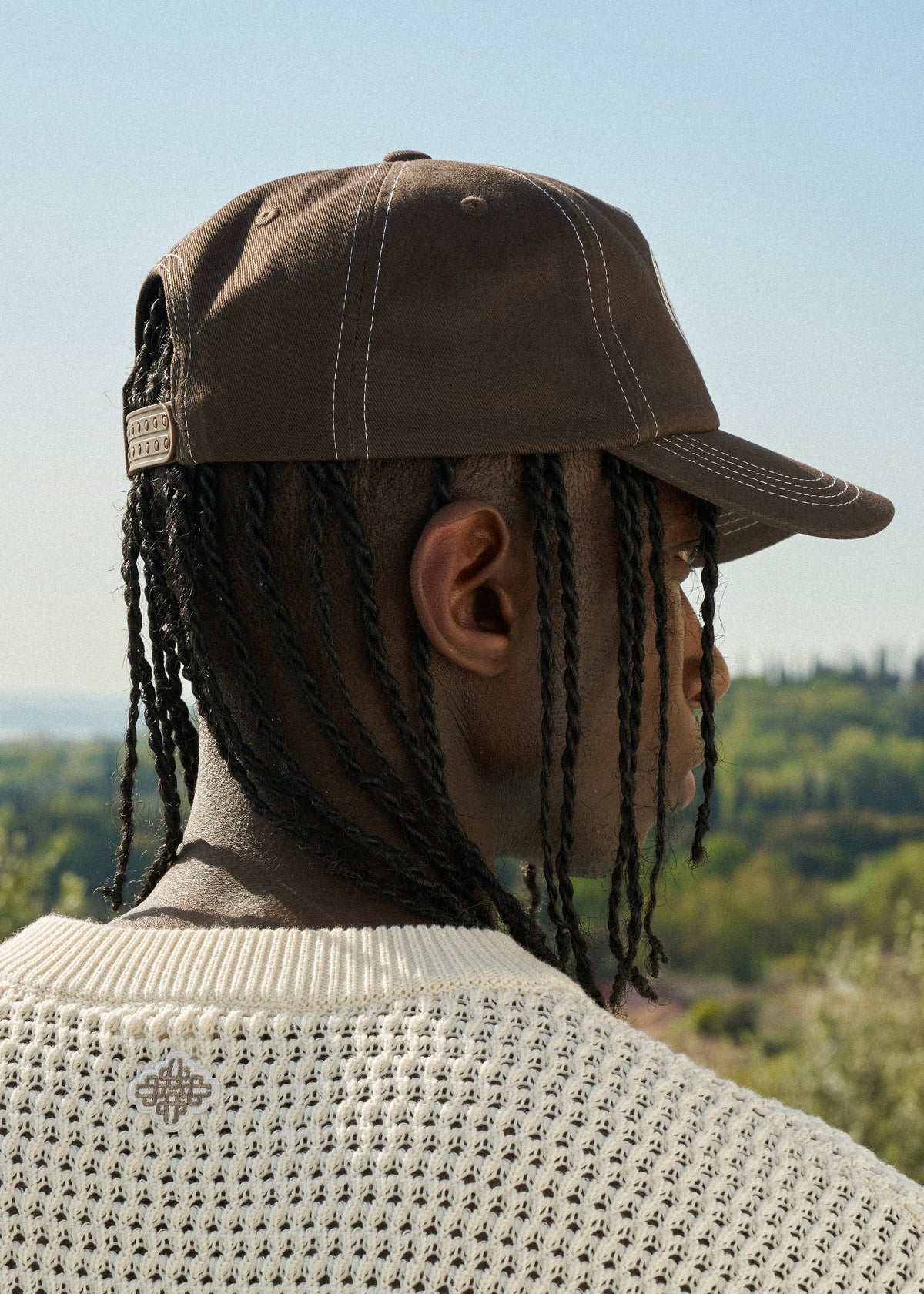 Person wearing a brown cap and light crochet knit top with a scenic background