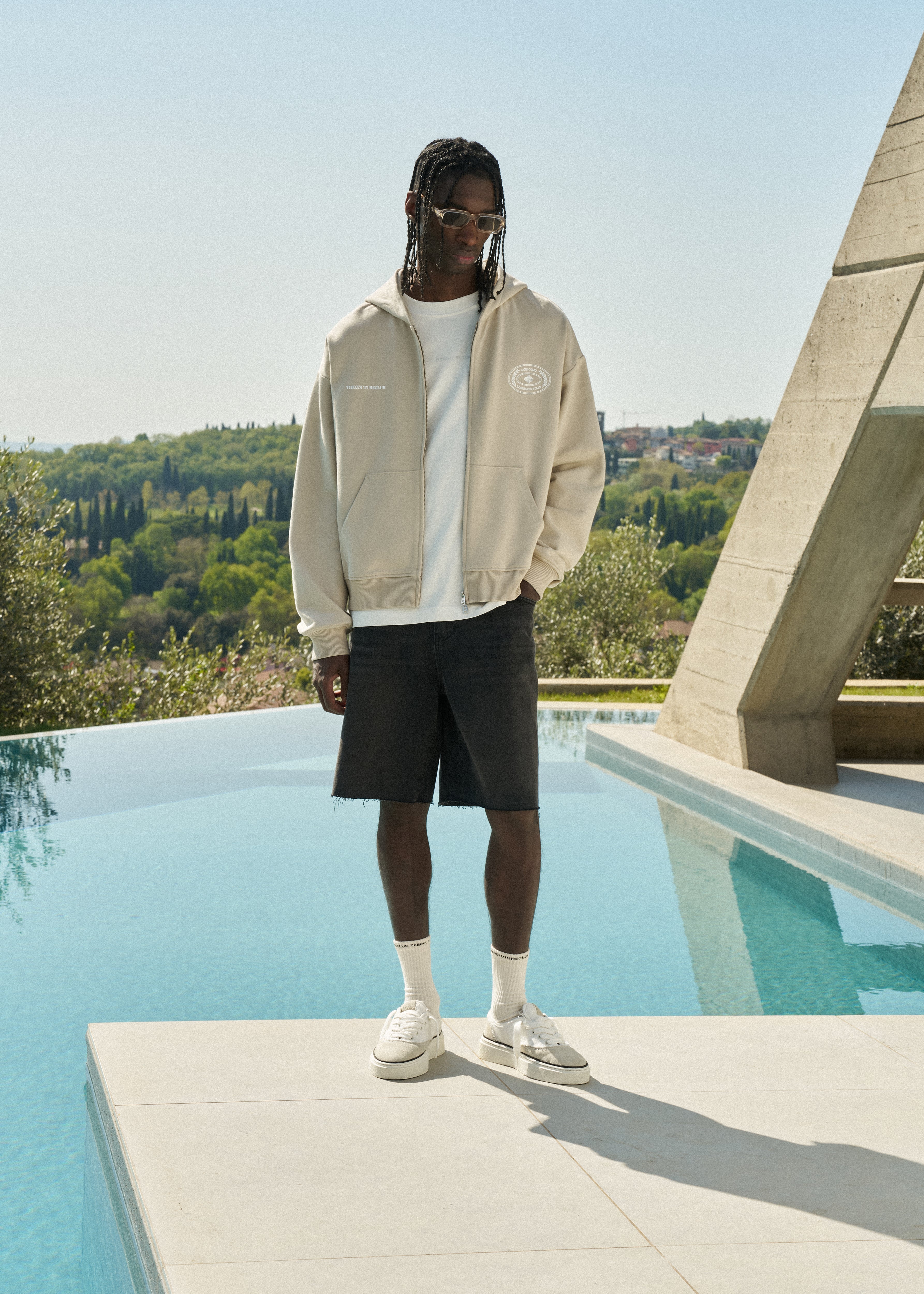 Man wearing a beige hoodie and black shorts standing by a pool with greenery in the background