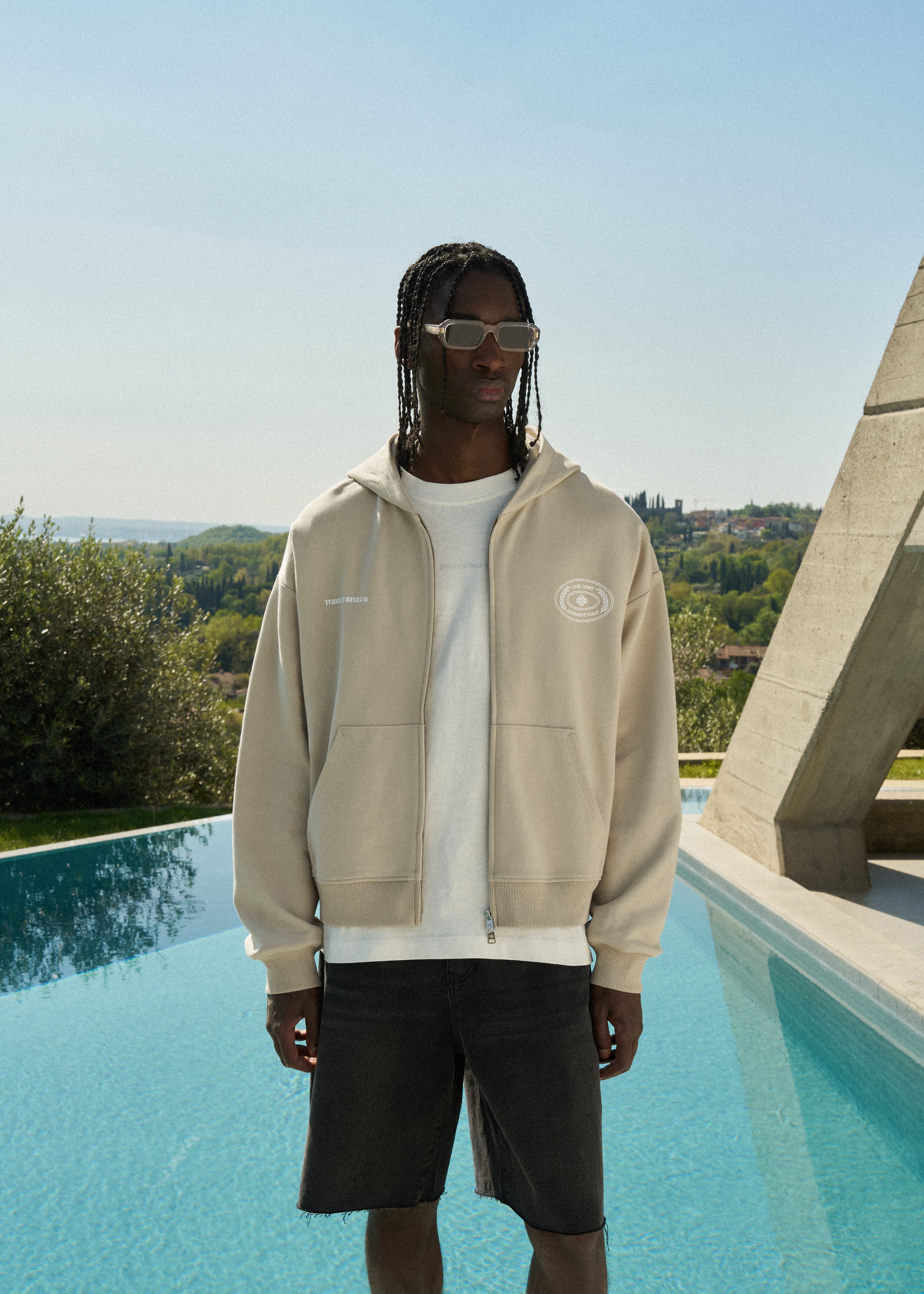 Man wearing a beige hoodie and sunglasses by a pool with a scenic background