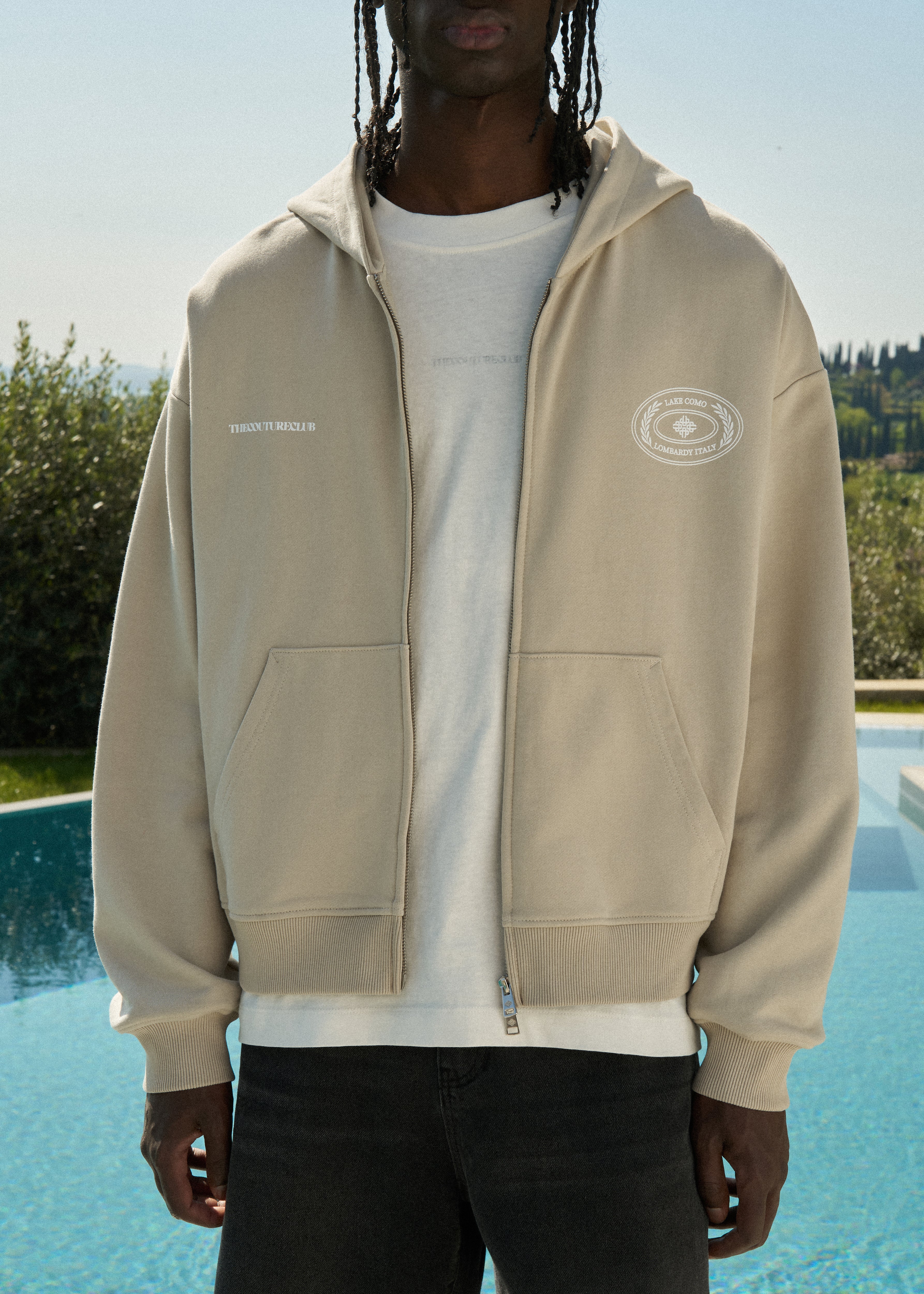 Man wearing a beige hoodie with brand logos by a poolside