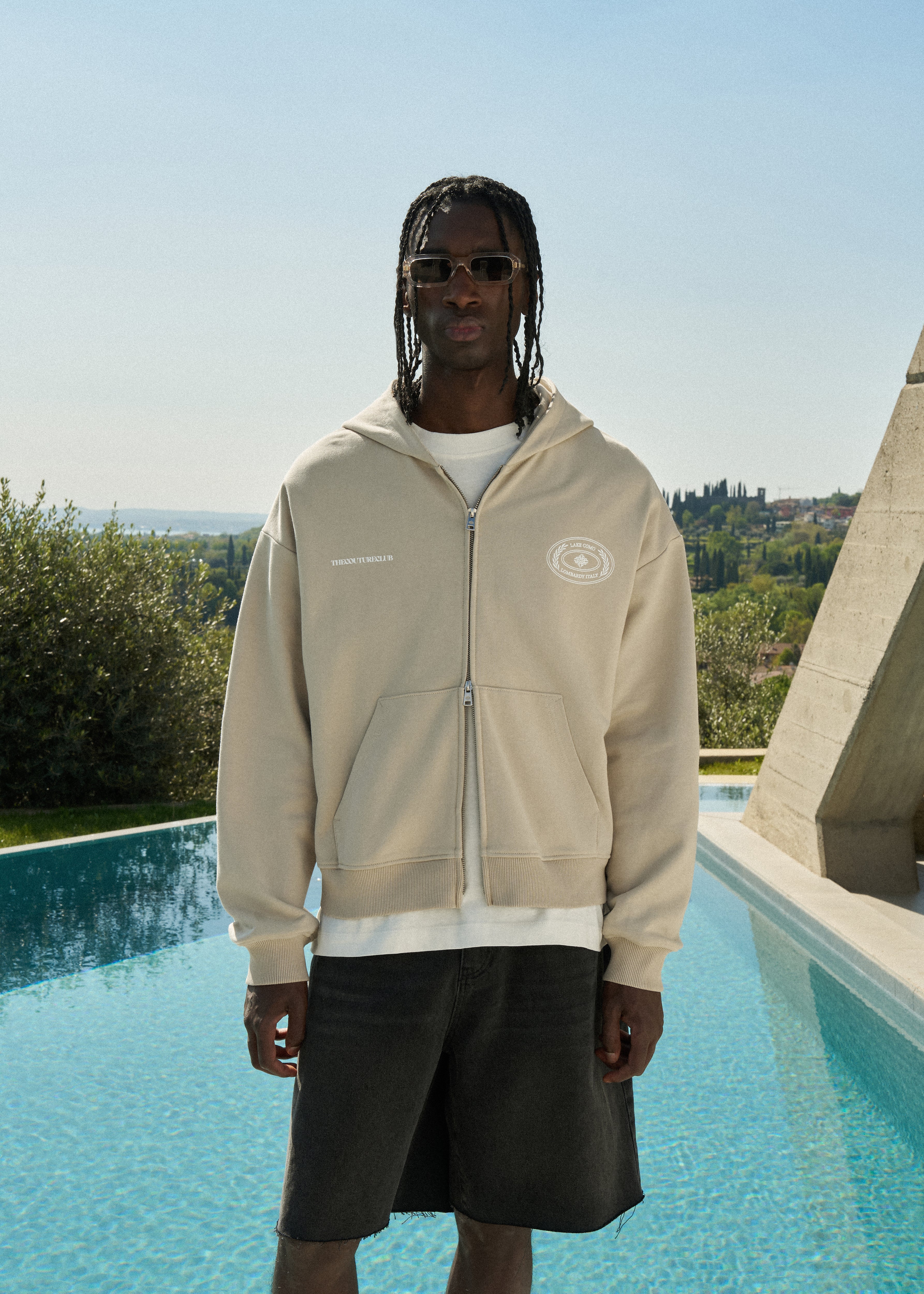Man wearing a beige hoodie with a logo, standing by a pool with a scenic background.
