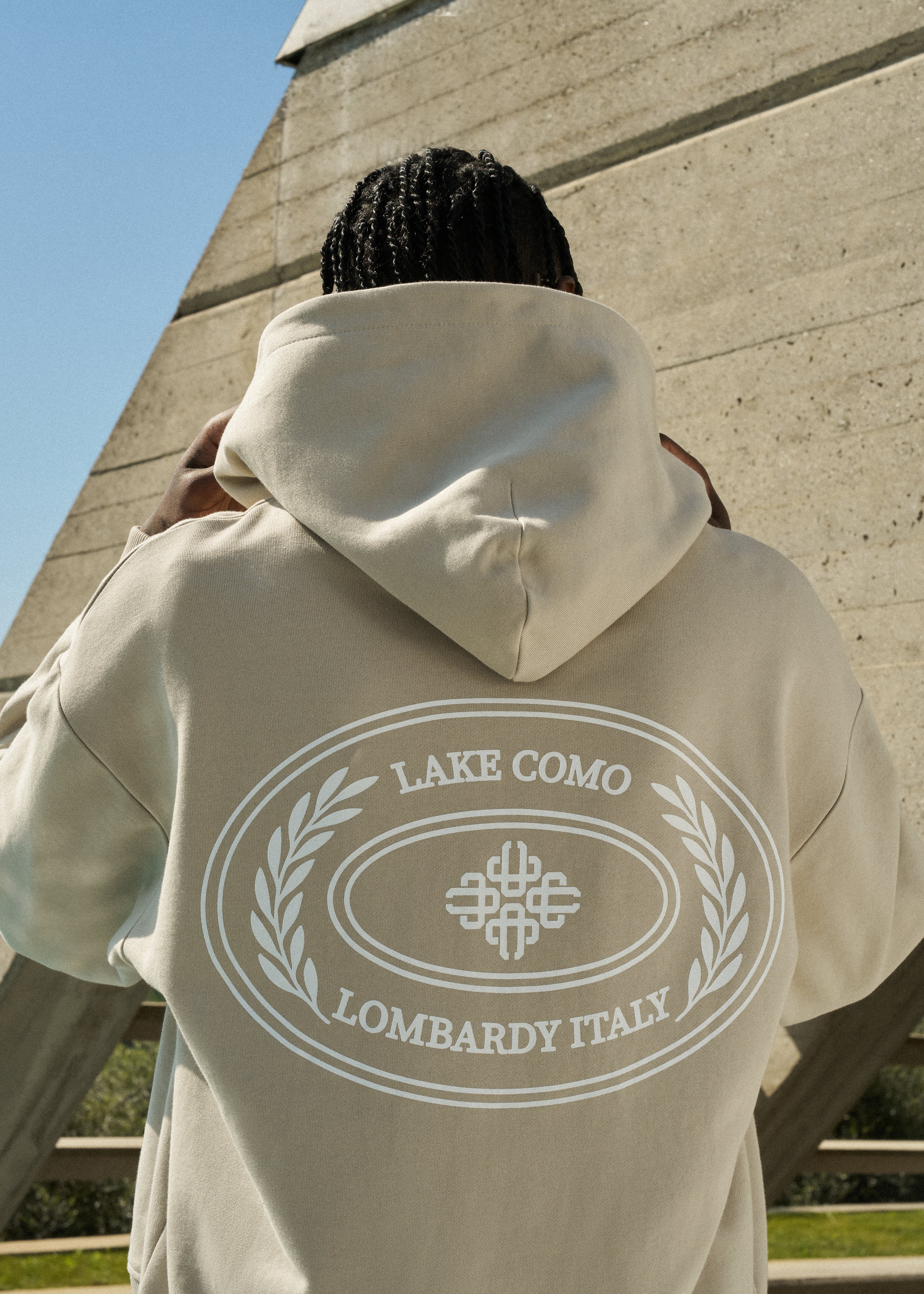Man wearing a beige hoodie with 'Lake Como Lombardy Italy' text on the back, standing against a concrete wall.