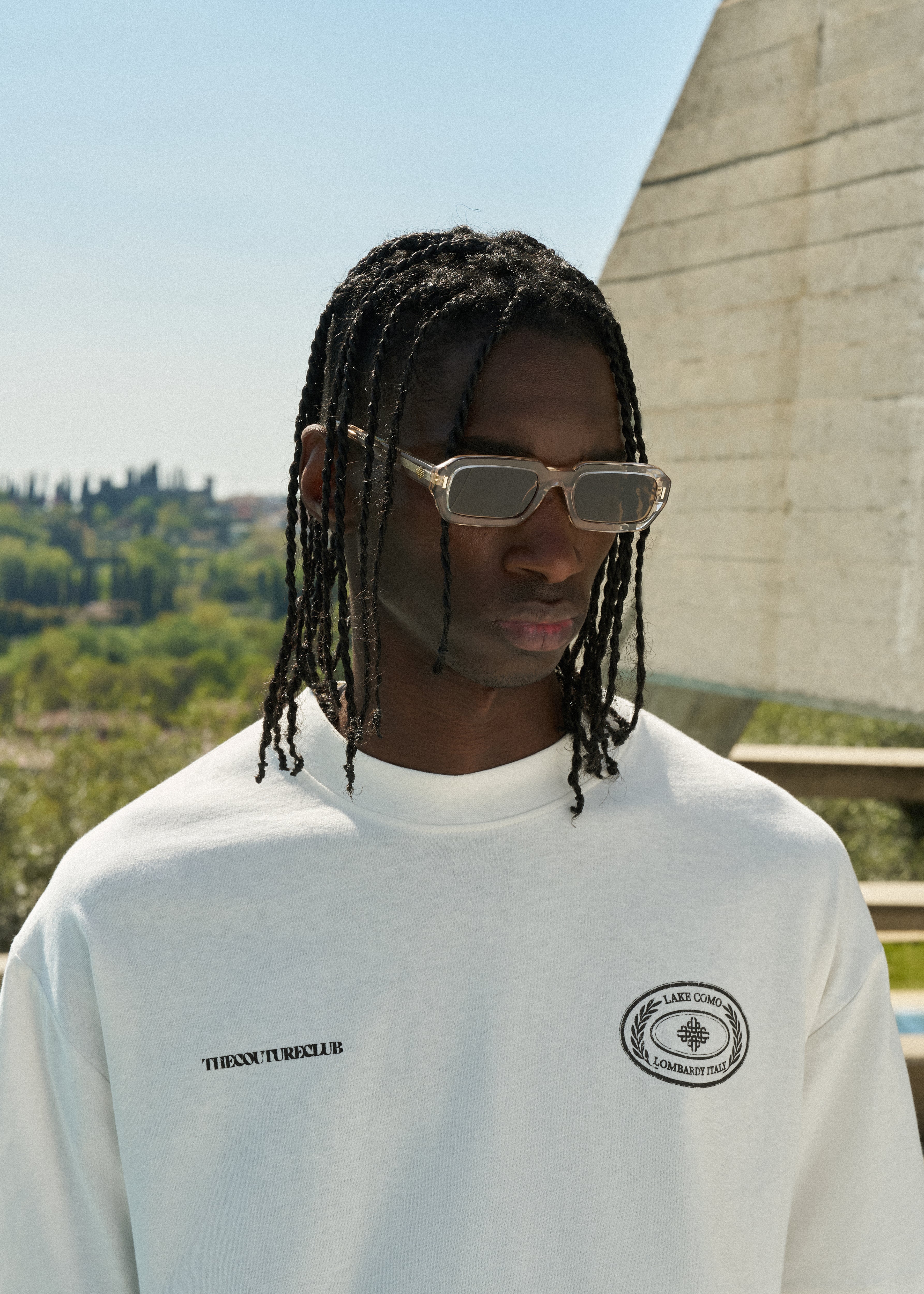 Man wearing a white t-shirt with logos, standing outdoors with greenery and a building in the background.