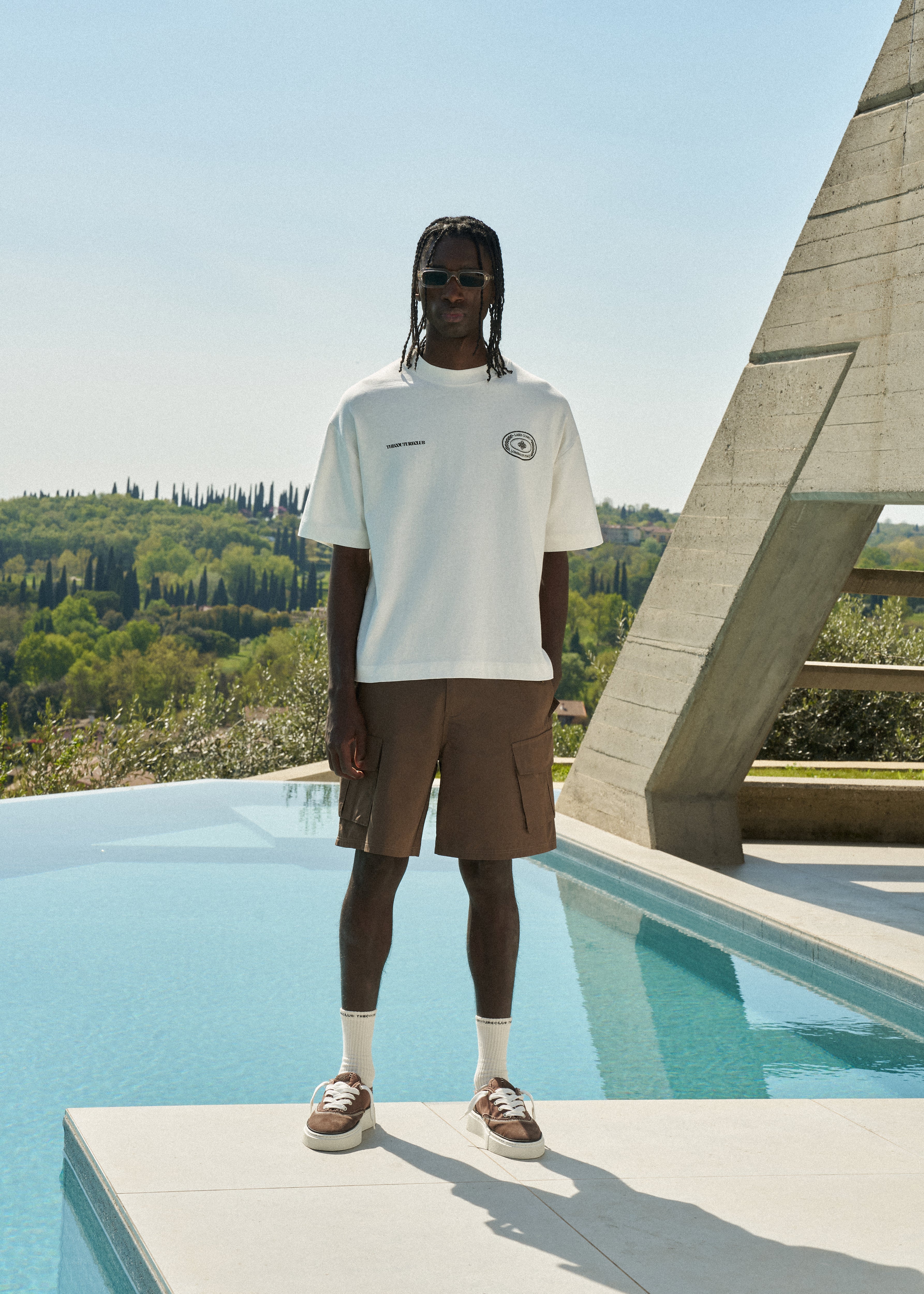Man wearing a white t-shirt and brown shorts standing by a pool with greenery in the background