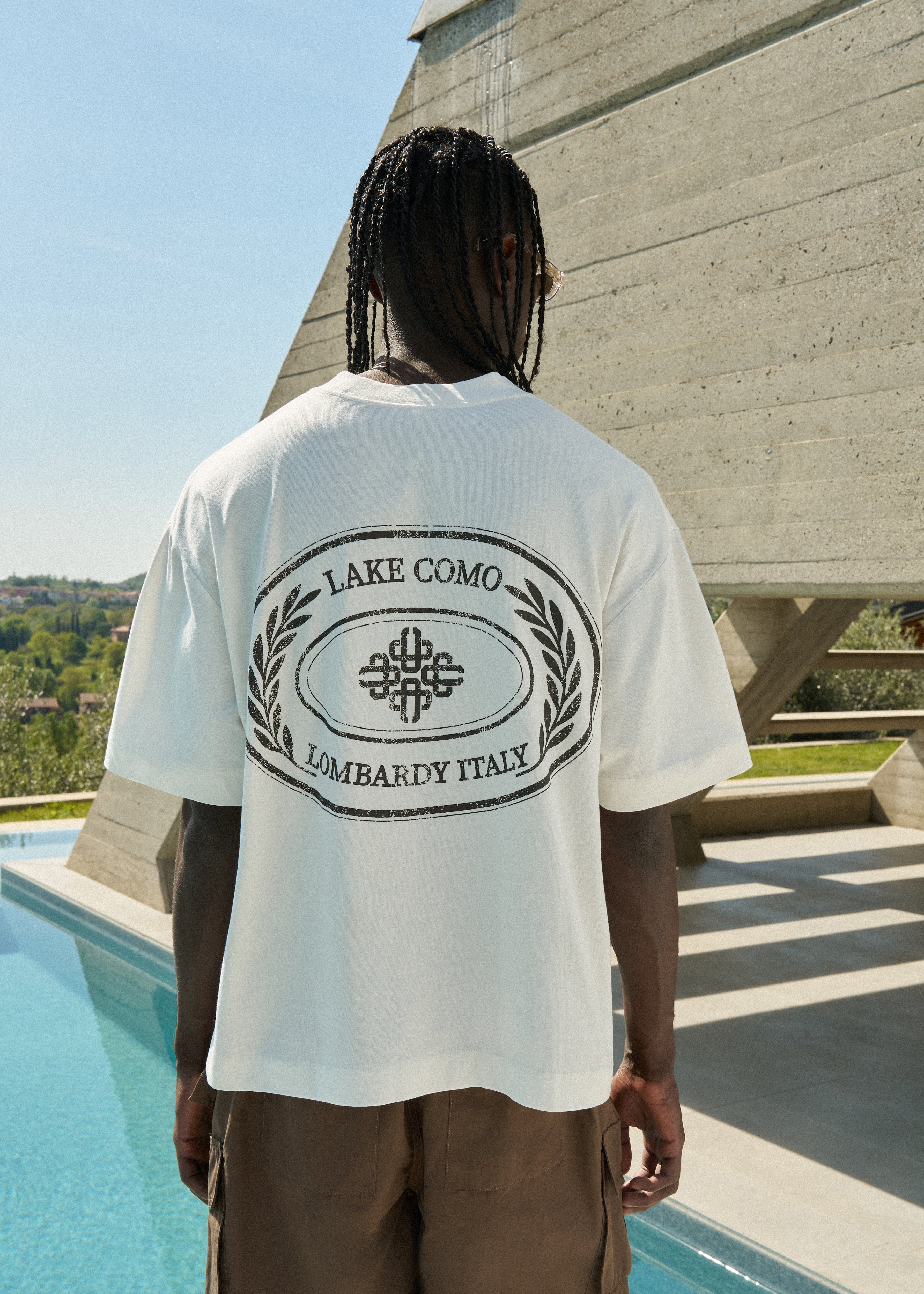 Man wearing a white t-shirt with 'Lake Como Lombardy Italy' design and sunglasses standing by a pool.